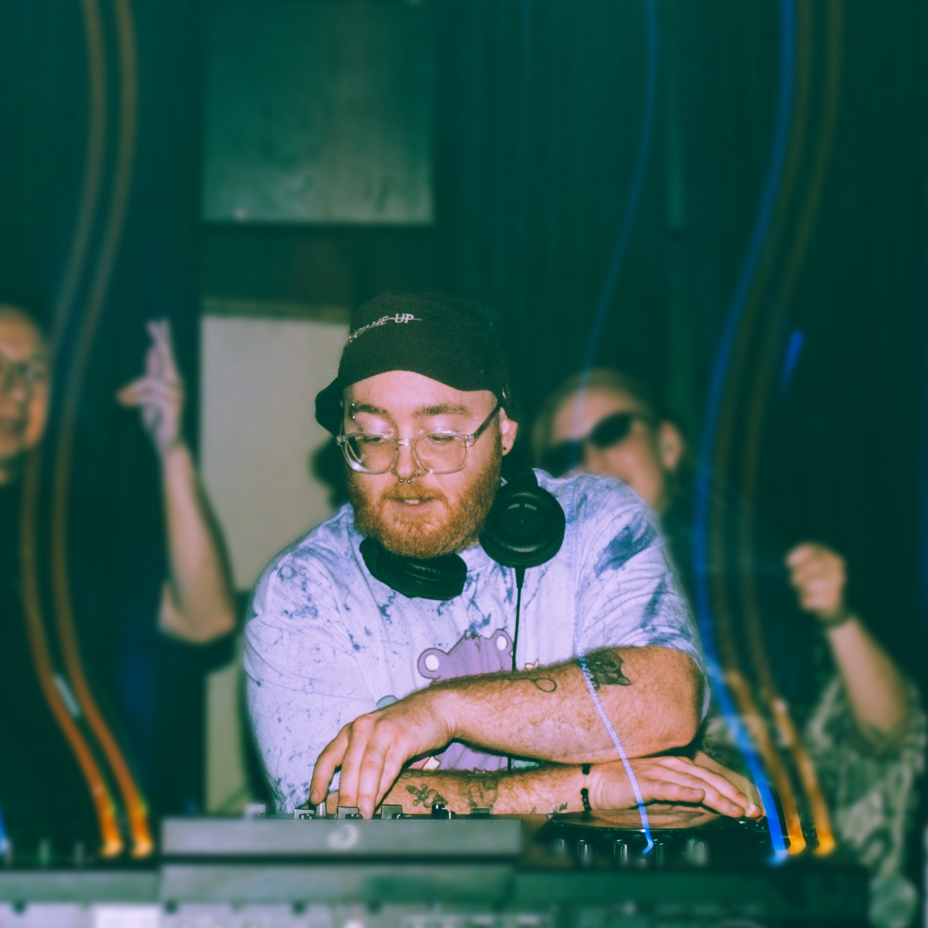 A person with glasses and facial piercings wearing a black beanie and headphones around their neck, seen through a vanity mirror. The person is wearing a light-colored T-shirt with a purple bear graphic. They are looking down at a turntable or DJ equipment, with other people in the background.