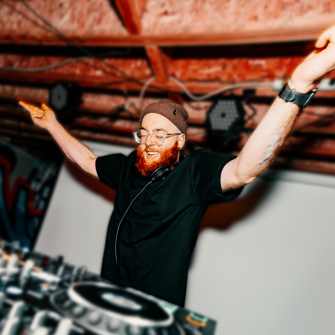 A DJ with red hair and beard, wearing glasses and a beanie, is celebrating with arms raised in a music studio.