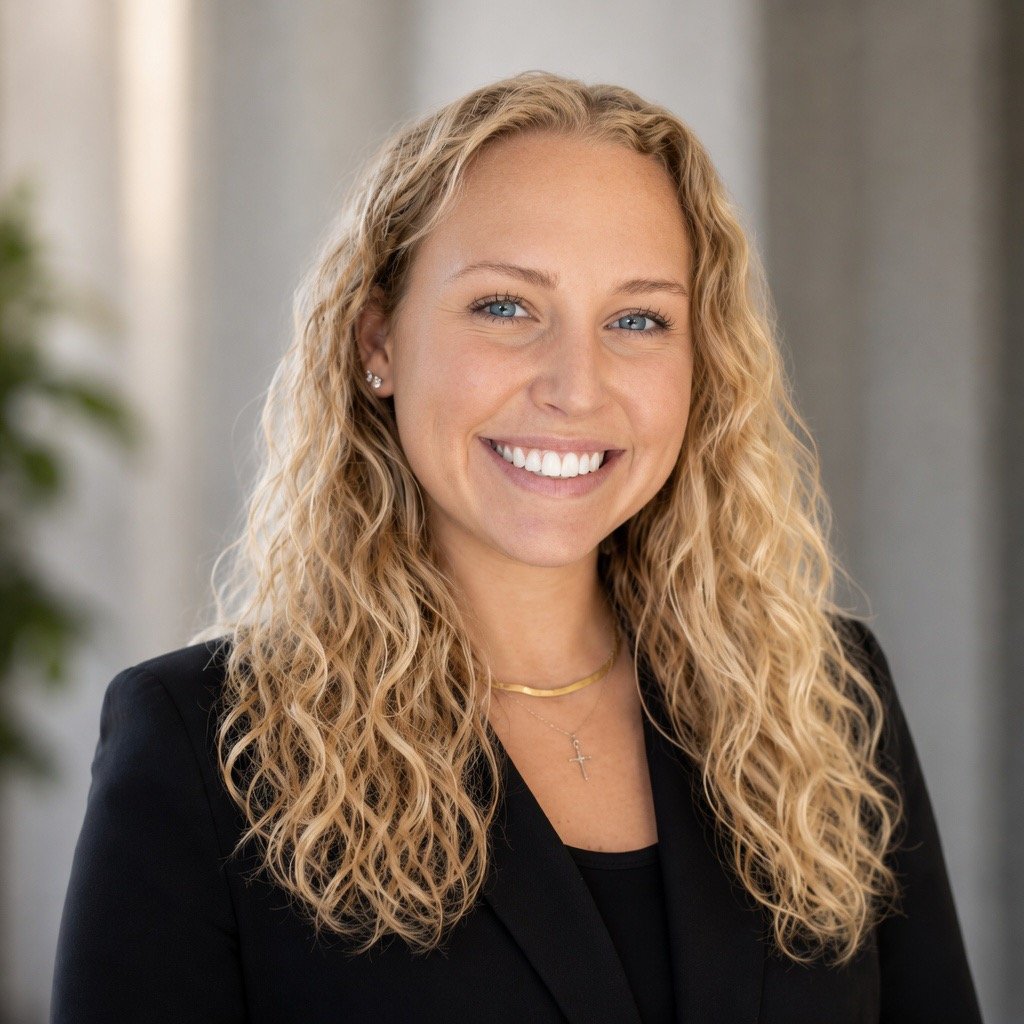 Professional headshot of a woman with curly blonde hair, blue eyes, and a bright smile, wearing a black blazer, gold necklace, and earrings, in a modern indoor setting.