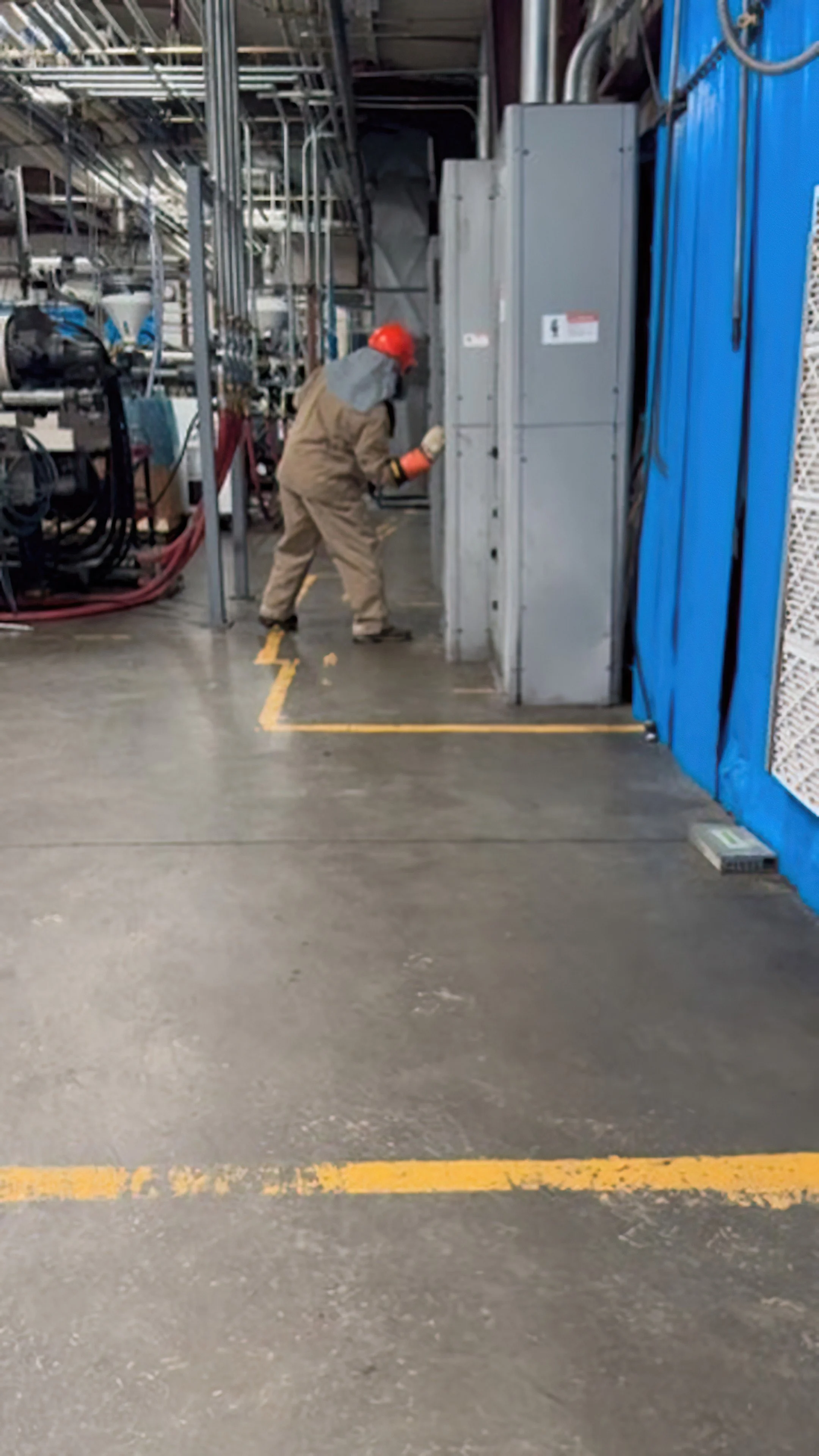 A worker wearing a red hard hat, brown coveralls, and gloves is inspecting electrical panels in an industrial setting with machinery, cables, and metal structures around.