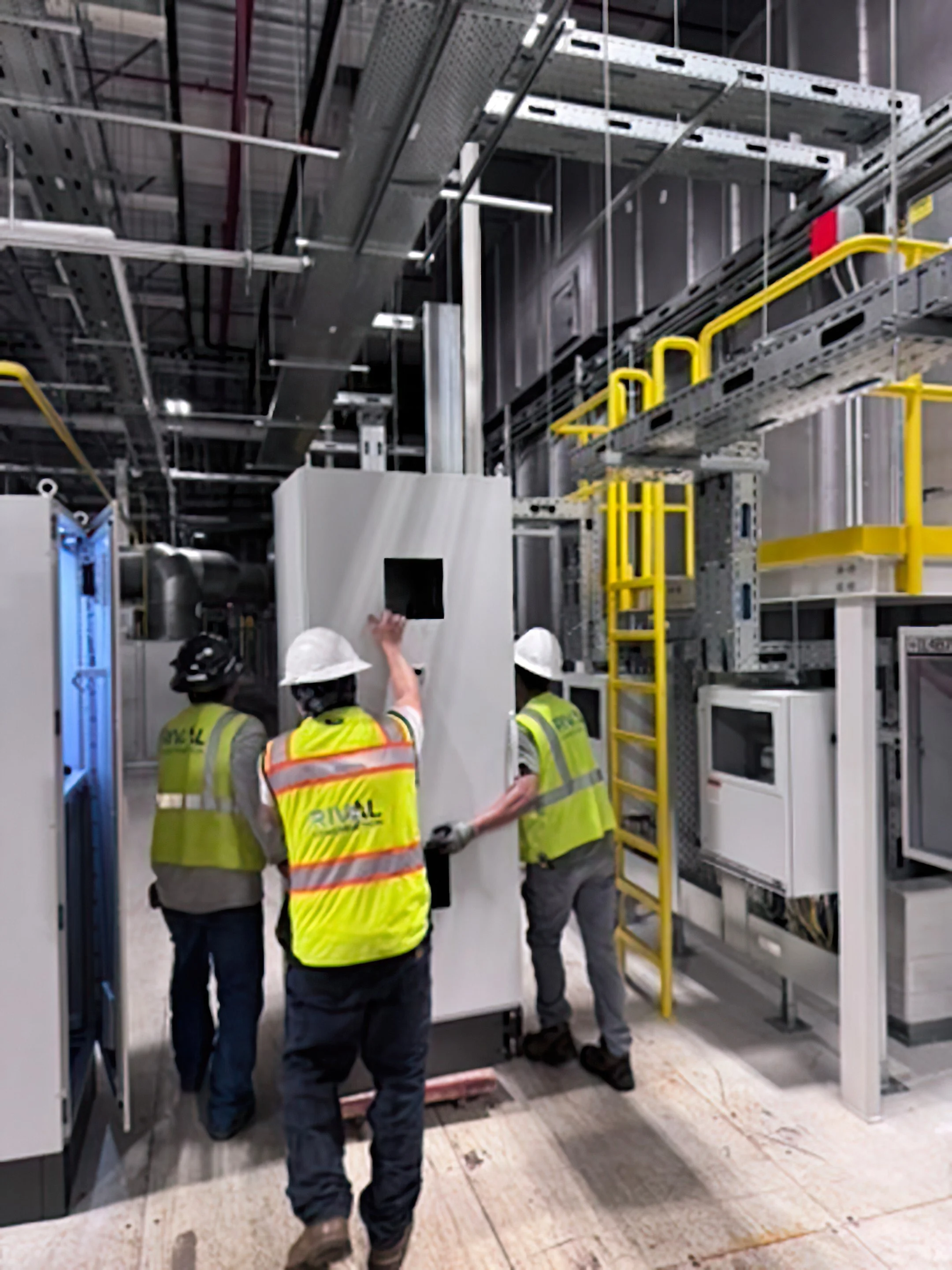 Three construction workers wearing yellow safety vests and white hard hats are working inside an industrial facility or data center with metal racks, scaffolding, and electrical wiring overhead.
