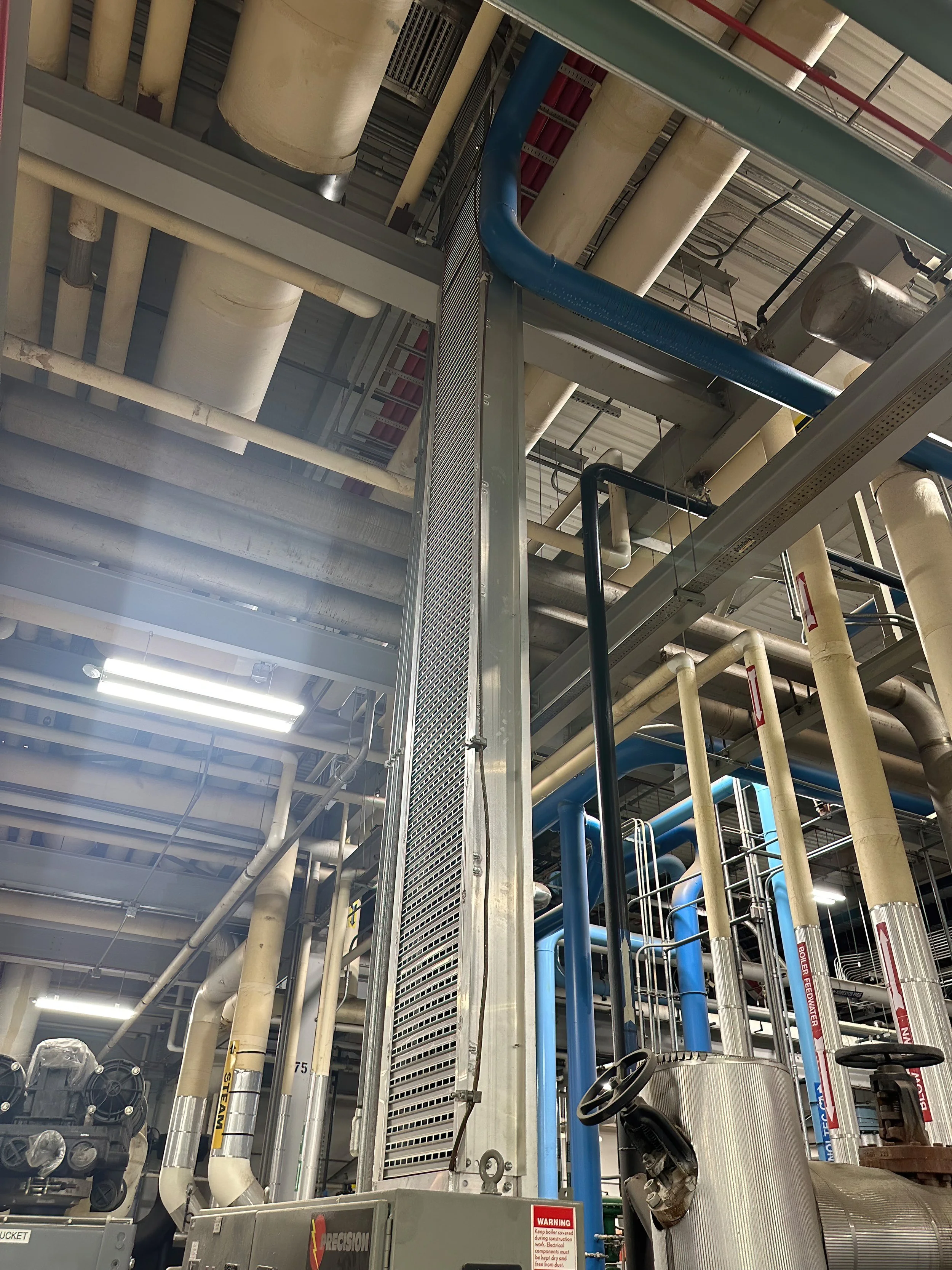 Industrial piping system with large beige pipes, blue pipes, metal grates, and safety warning signs in a facility.