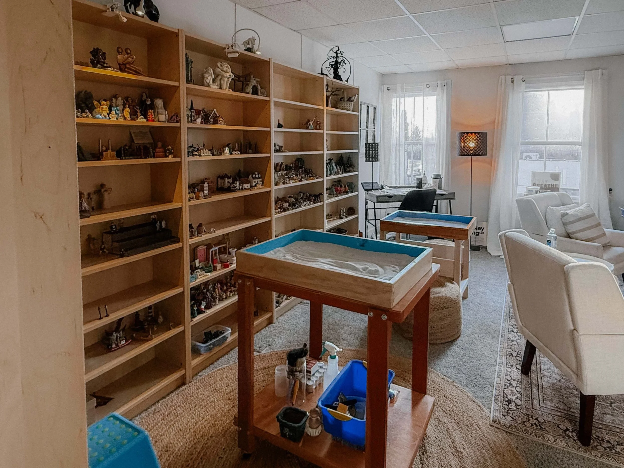 A room with wooden bookshelves filled with figurines and collectibles, a desk with a chair, and a comfortable armchair near windows with white curtains. There are cleaning supplies, containers, and small items on the floor and table.