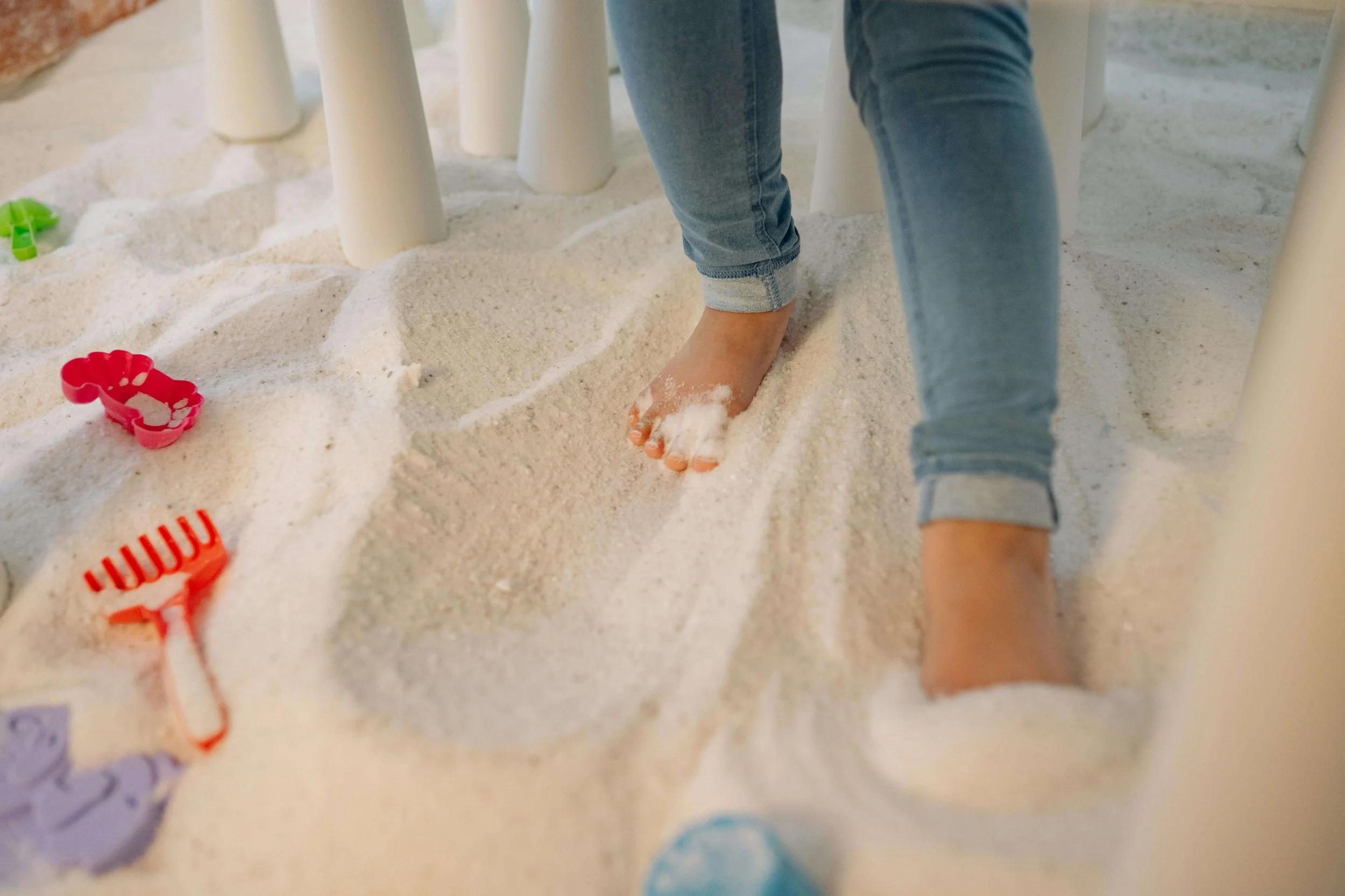 Child's bare foot and leg in blue jeans walking in white sand under a table with white legs, with colorful sand toys and a plastic rake scattered around.