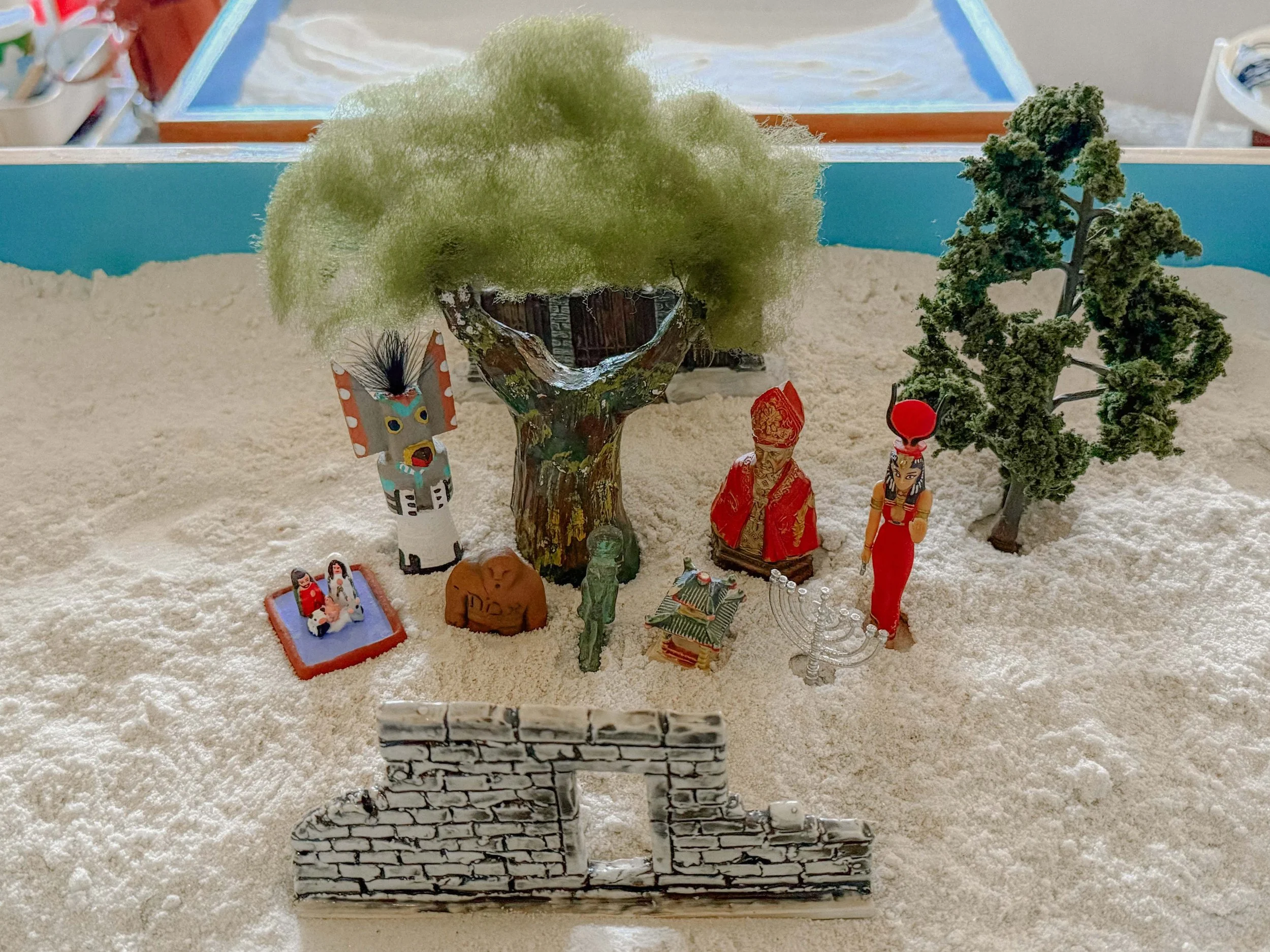 Miniature scene with trees, figurines, and decorative items on a sandy surface, enclosed by a low brick wall at the front.