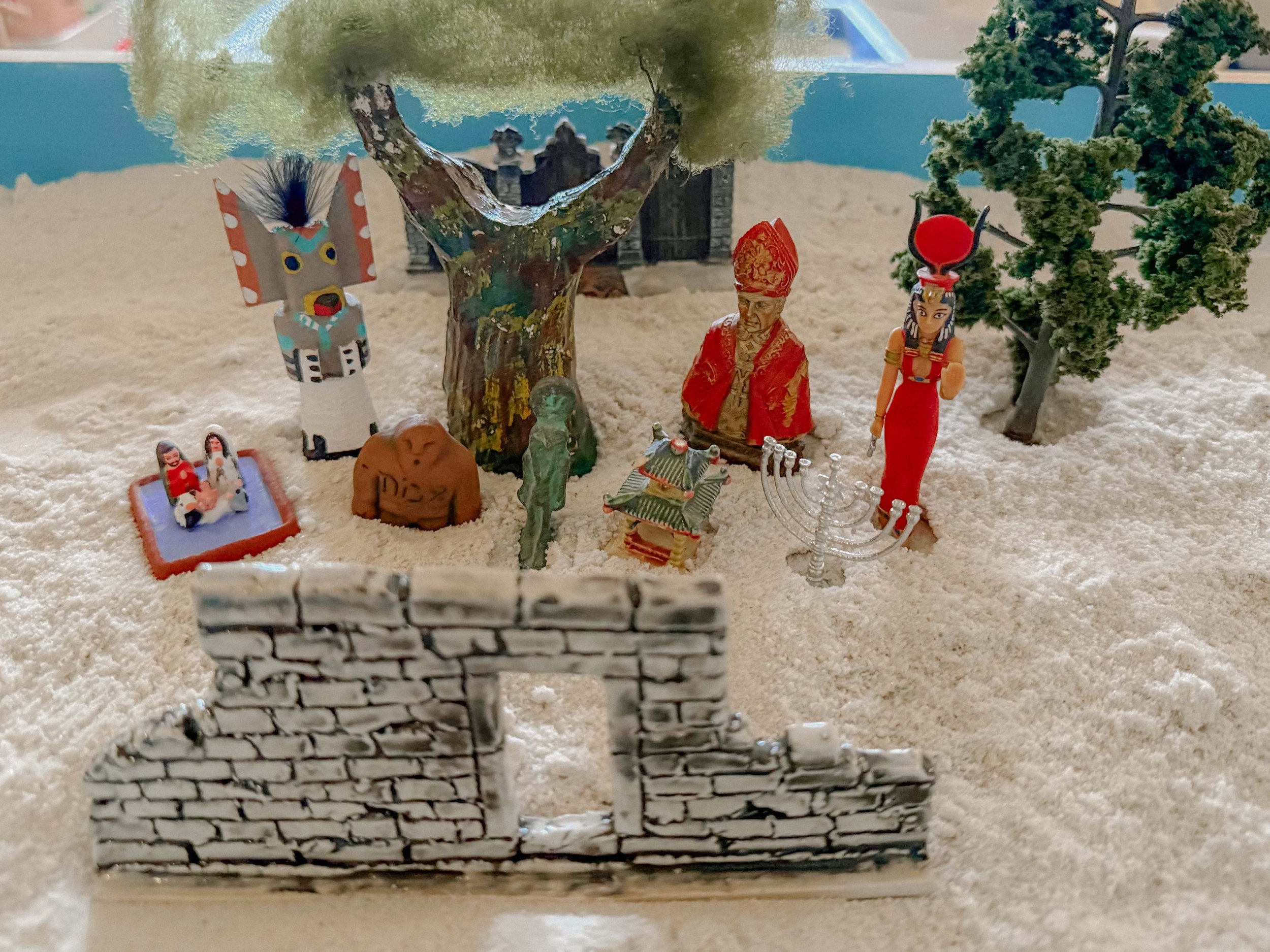 Miniature scene with figurines, trees, and a small fence on a textured surface.