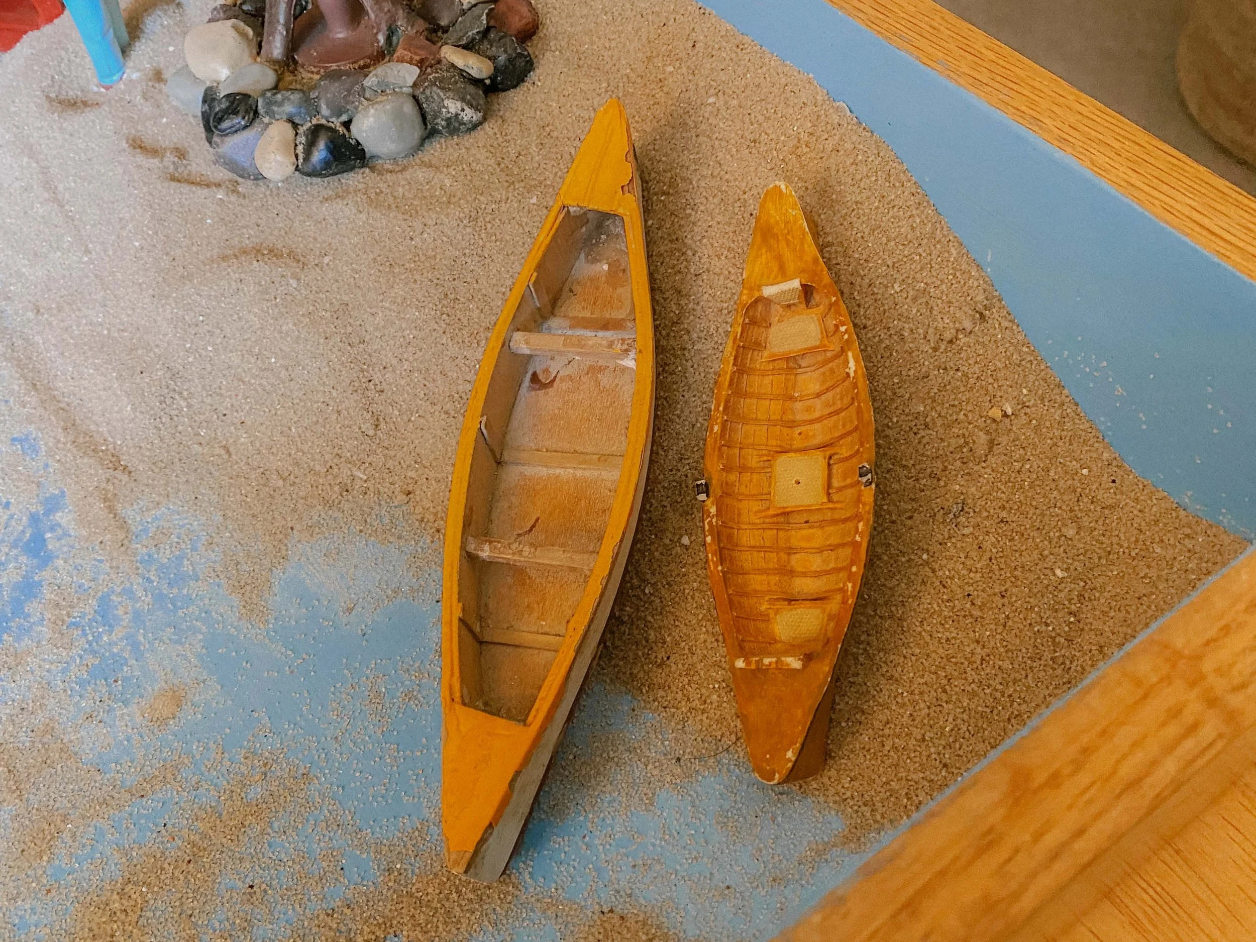 An empty yellow canoe and a fishing boat are placed on a sandy beach with some rocks near the shoreline and a blue wall in the background.