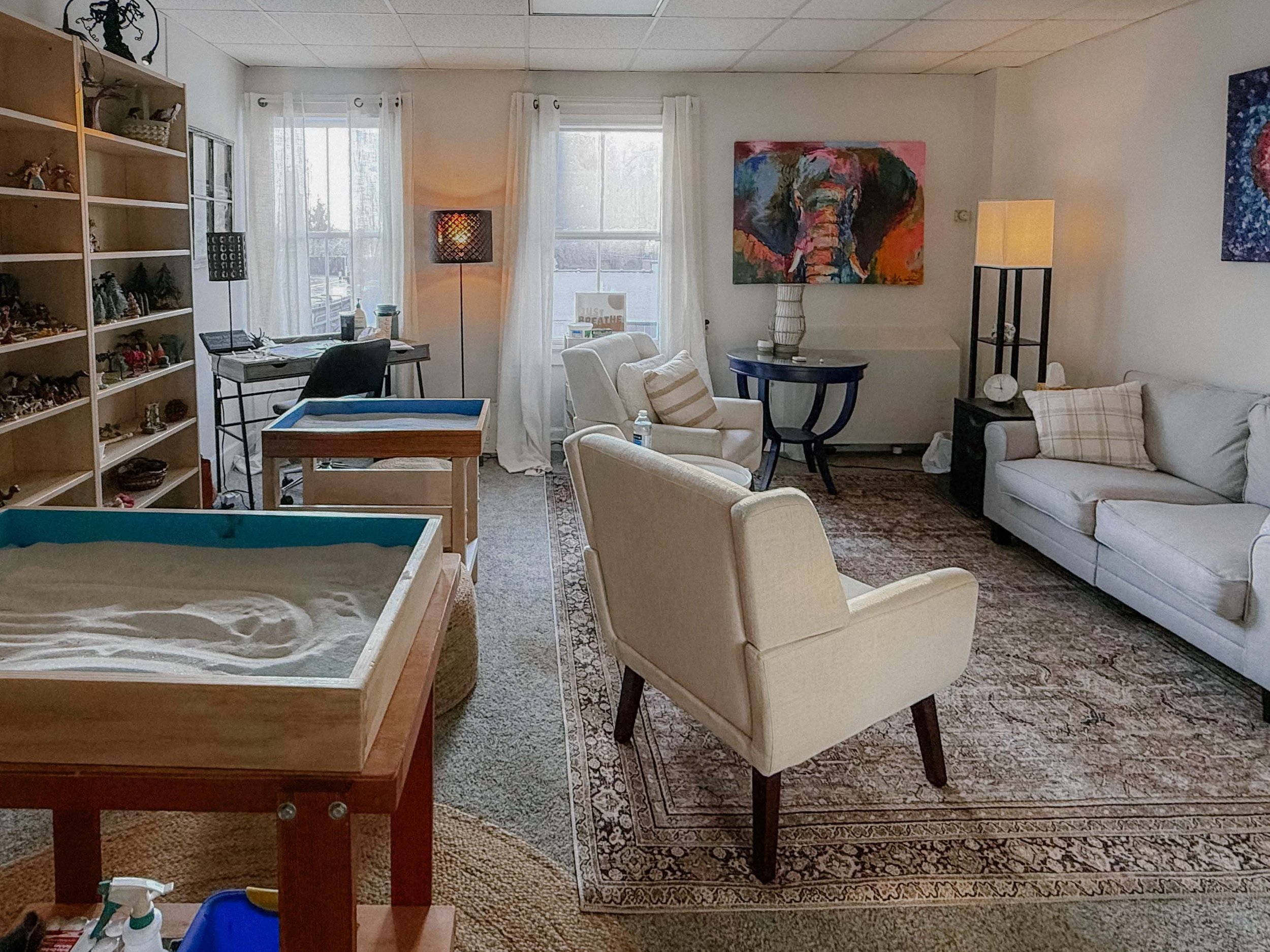 Living room with white curtains, artwork featuring an elephant, seating area with armchairs and a sofa, alongside a bookshelf filled with various items.