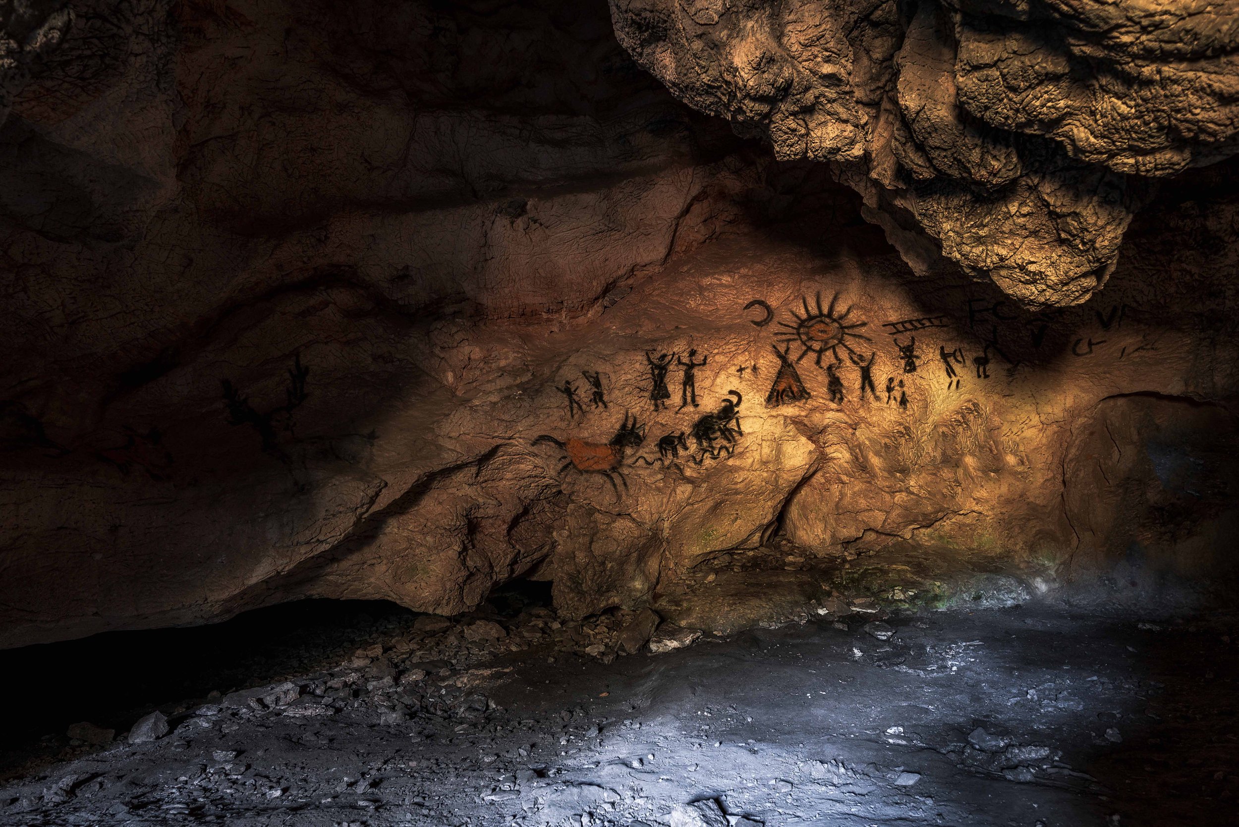 A dark cave interior with rocky walls and ceiling, featuring ancient cave paintings of human figures, animals, and symbols illuminated by subtle lighting.