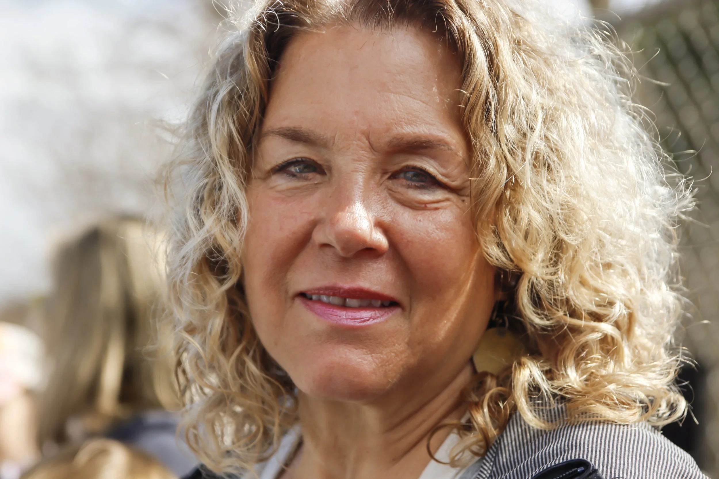 Close-up of a smiling woman with curly blonde hair and blue eyes outdoors.