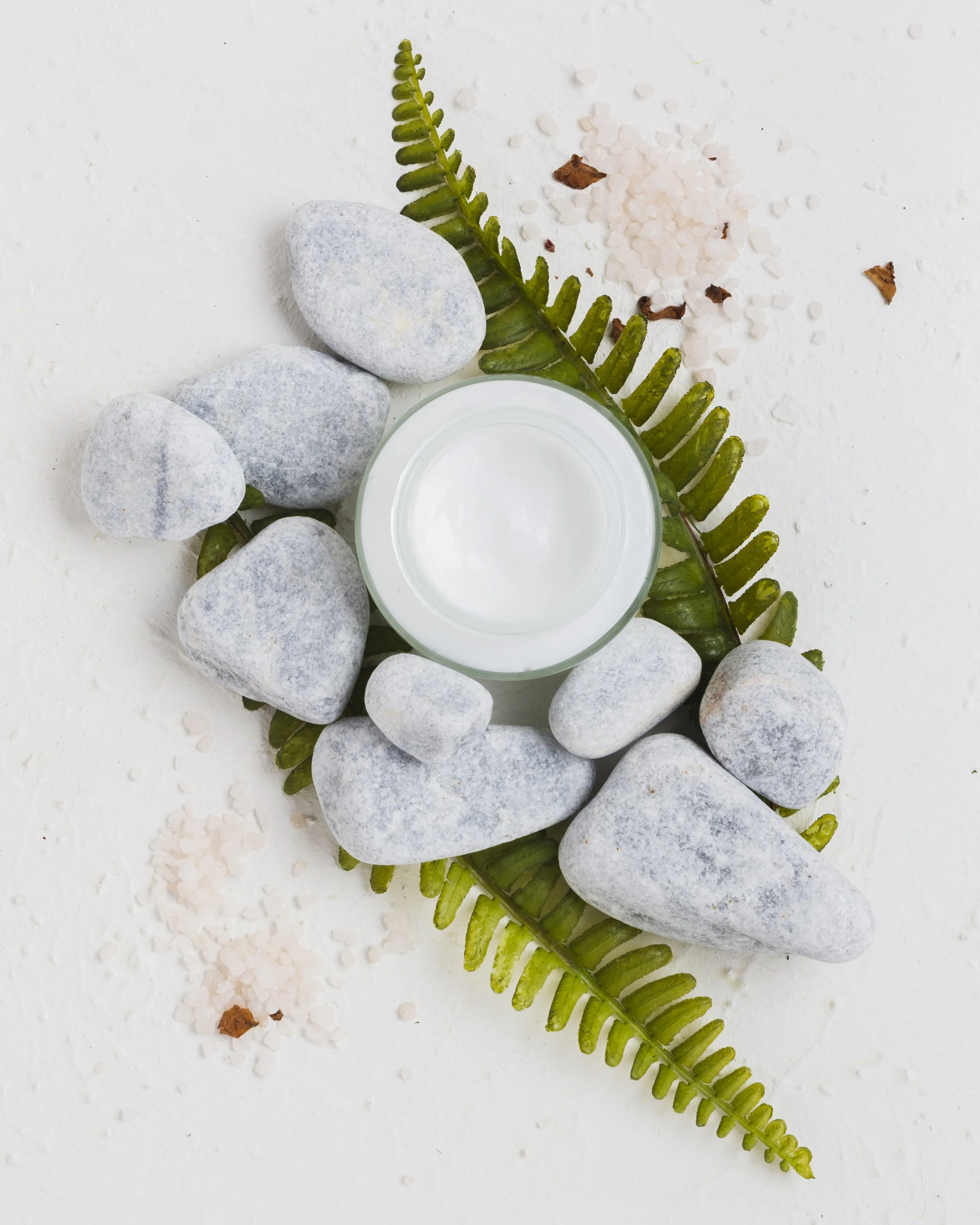 Des pierres grises, une branche de fougère verte, du sel rose de l'Himalaya et quelques feuilles de laurier sur un fond blanc