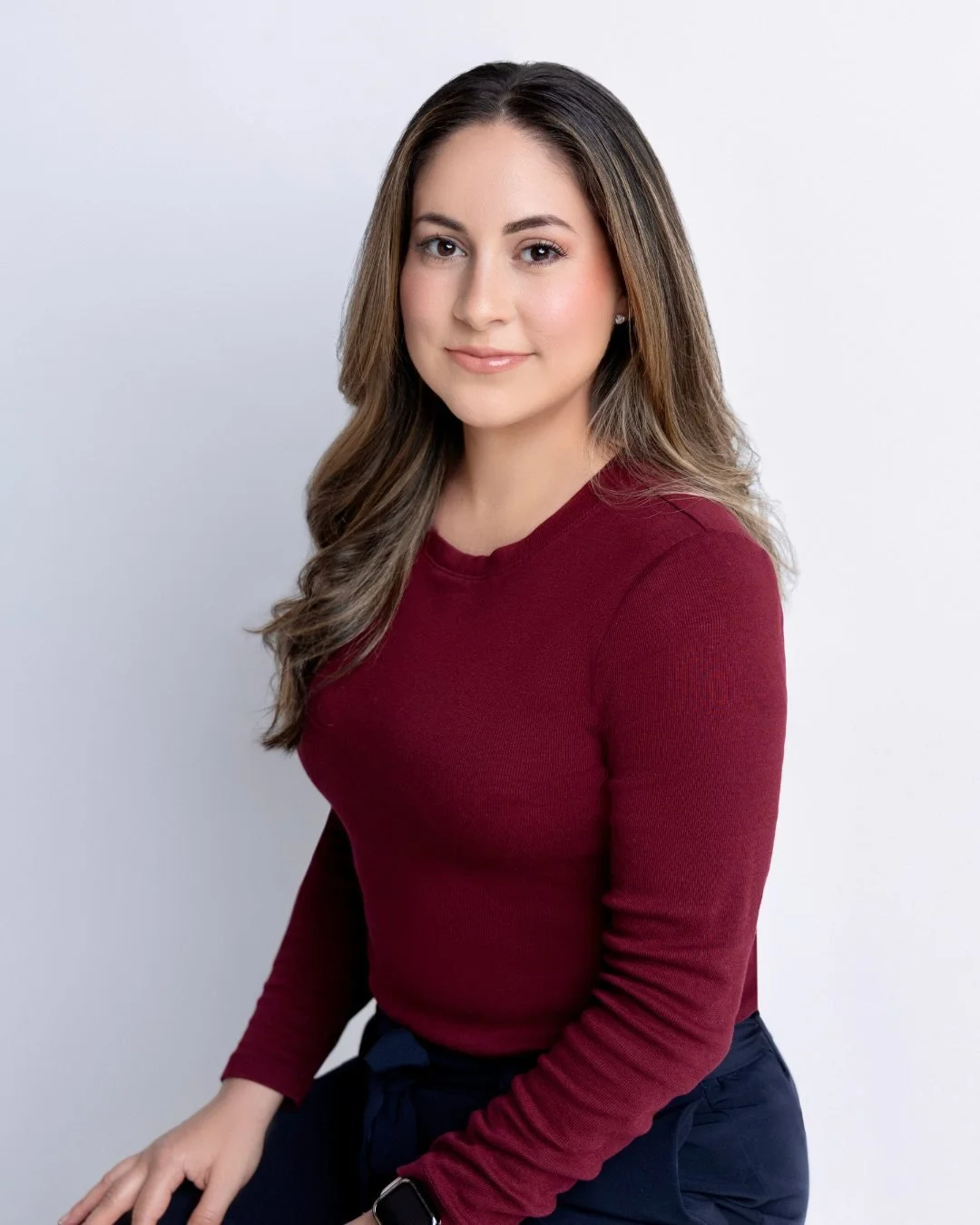 A young woman with long, wavy hair wearing a burgundy long-sleeved top and navy pants, sitting against a plain white background.