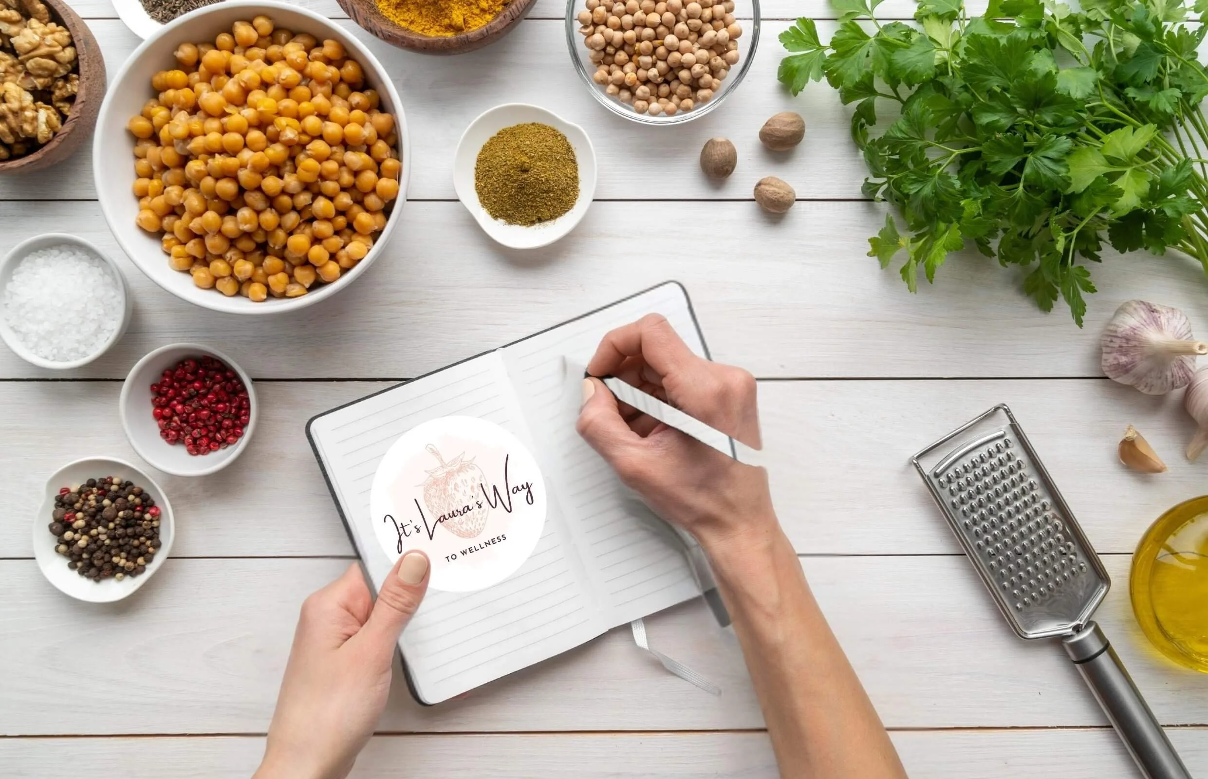 Overhead shot of a person writing in a notebook surrounded by bowls of spices, herbs, garlic, and a grater on a white wooden table, with parsley and oil on the side.