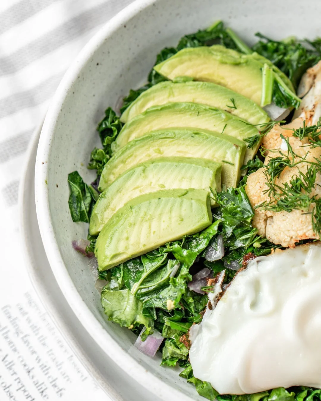 Close-up of a salad bowl containing sliced avocado, chopped kale, and a poached egg garnished with herbs.