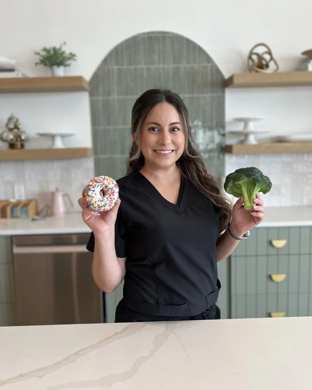 Woman in black scrubs smiling and holding a donut with colorful sprinkles in one hand and a head of broccoli in the other, in a modern kitchen.