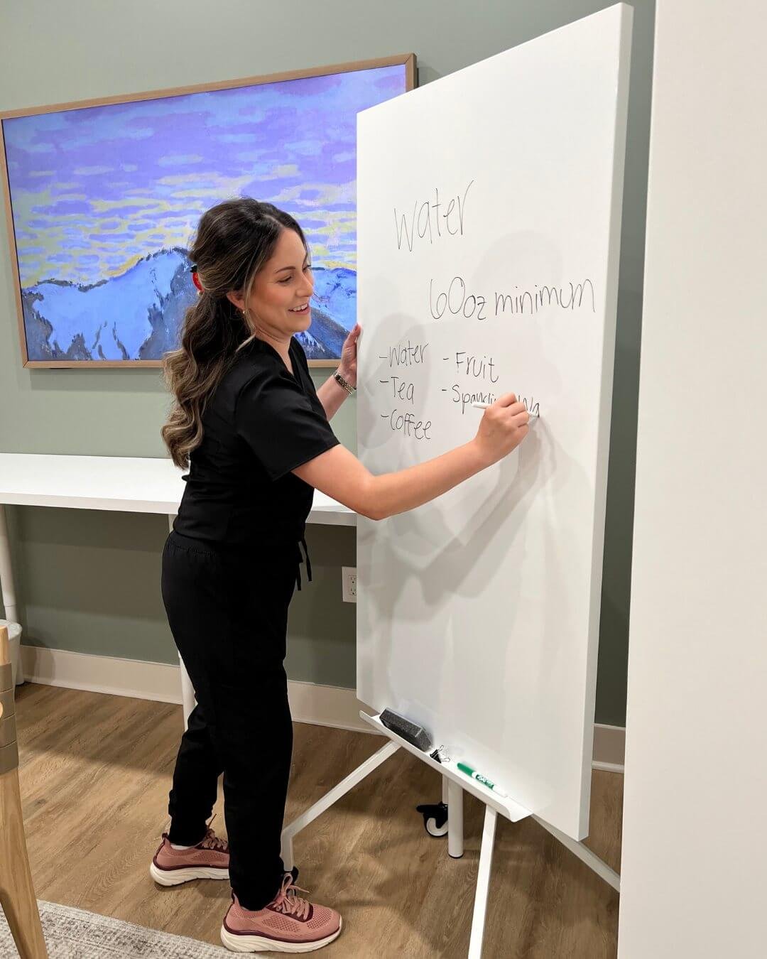 A woman writing on a whiteboard with a list of items including water, fruit, tea, sparkling water, and coffee, in a room with a wall-mounted TV showing a painting of mountains and water.