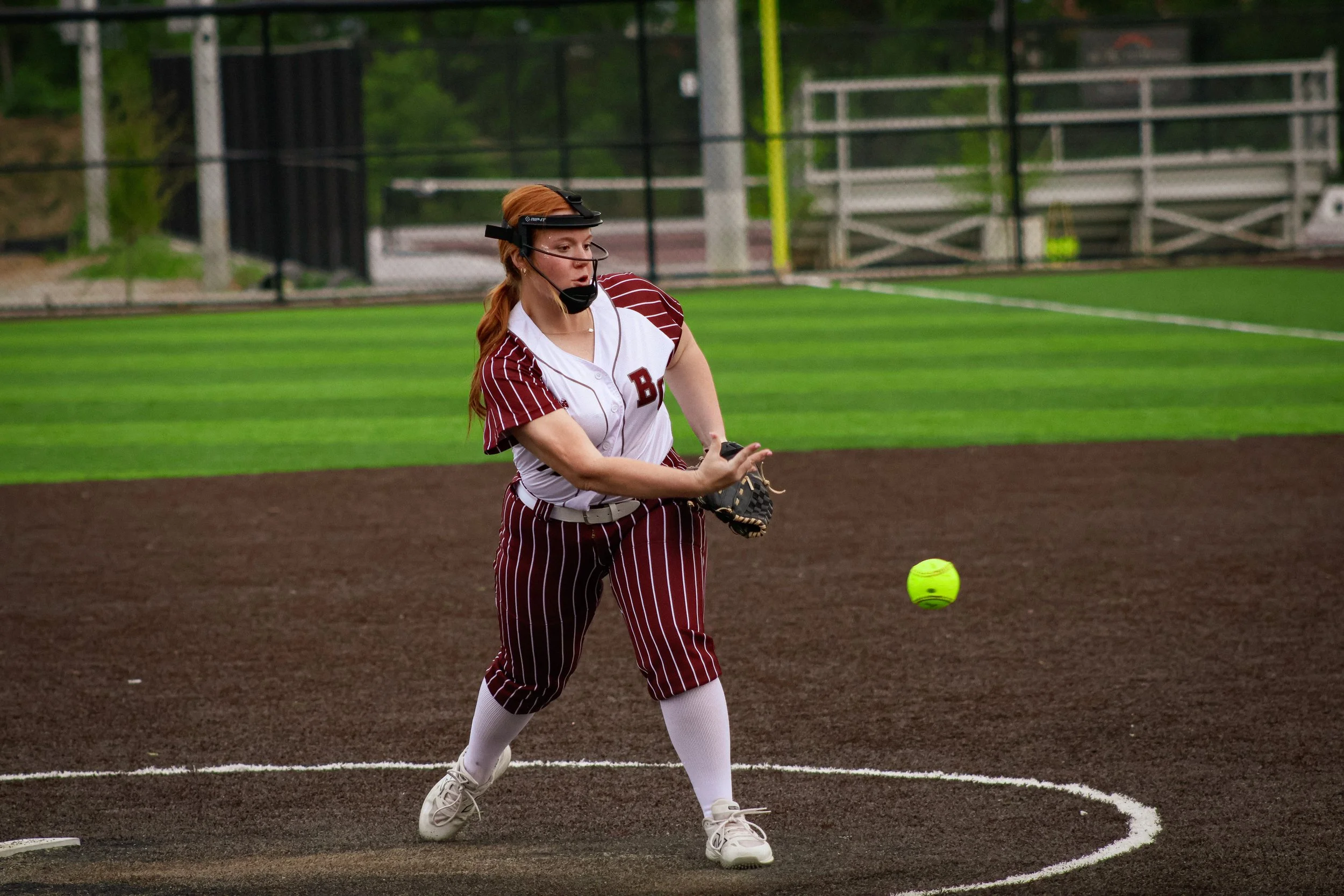 A female softball player with red hair, wearing a pinstriped uniform and a protective face mask, is standing on the field preparing to catch a yellow softball.