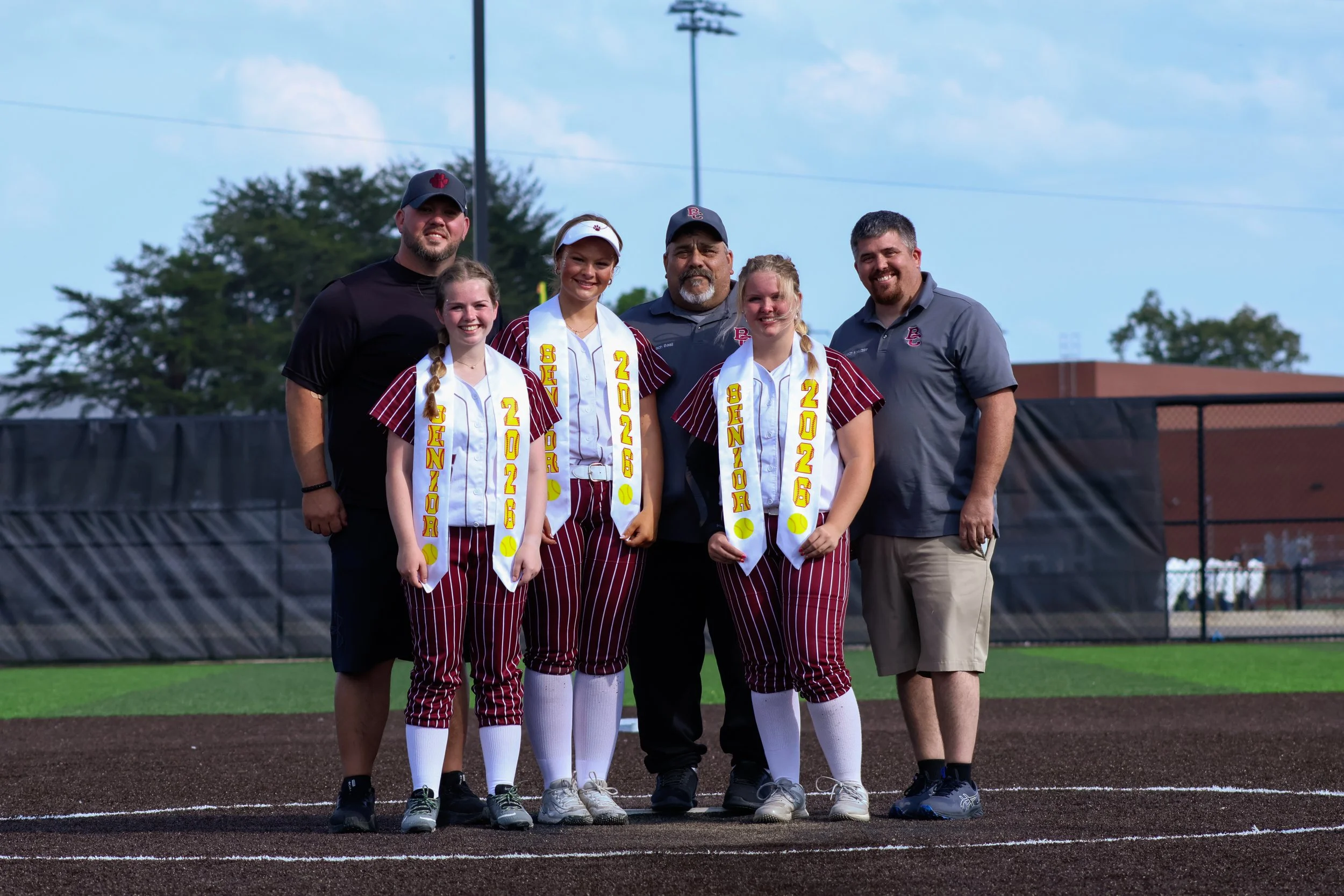 A group of six people, including three young female softball players in uniform, two adult males, and one older male, standing on a softball field with a blue sky and trees in the background. The players have medals and sashes that say 'Senior 2026'.