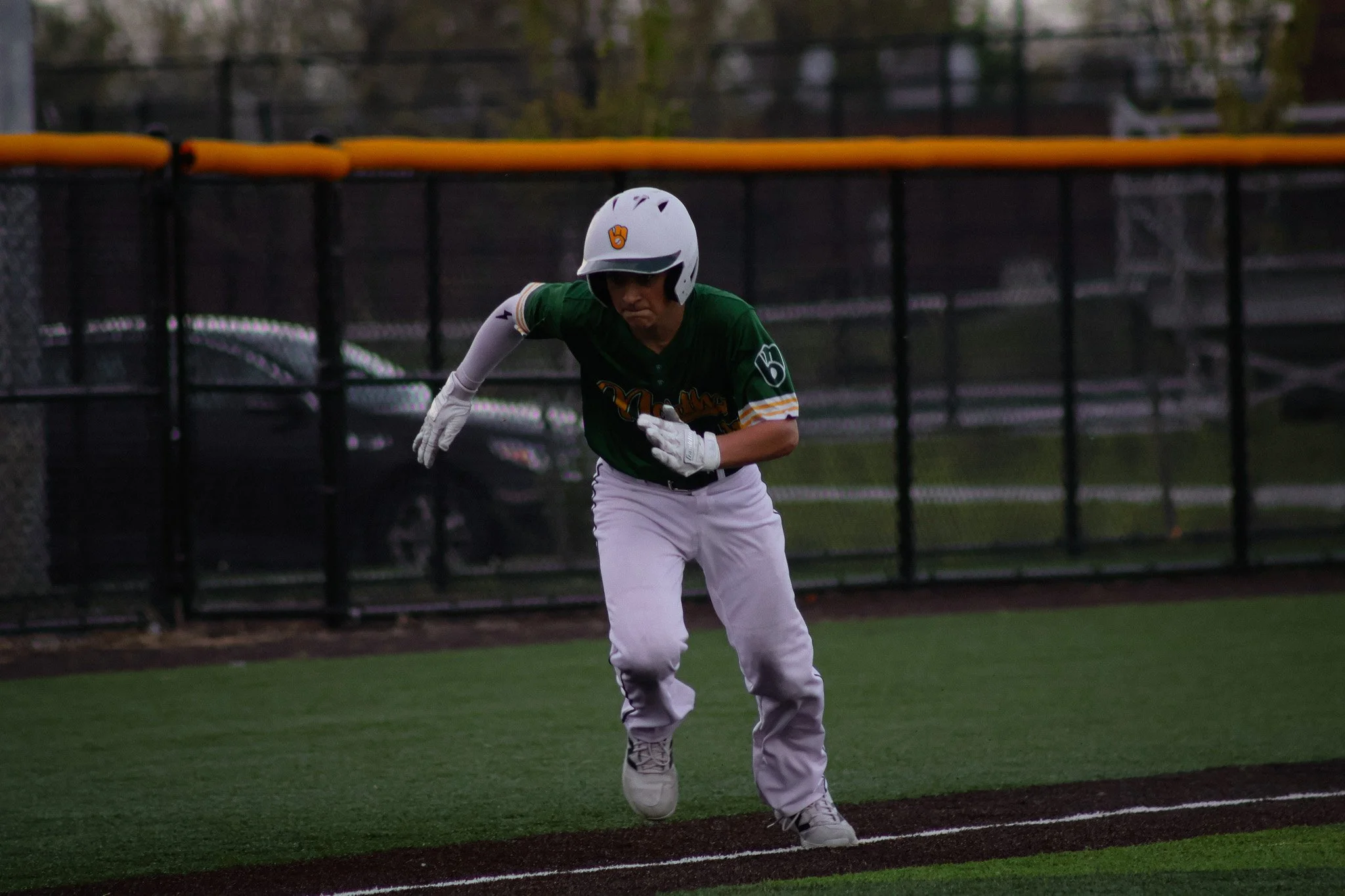 Baseball player in green jersey and white pants running on the field near the foul line, wearing a white helmet, with a black fence and trees in the background.