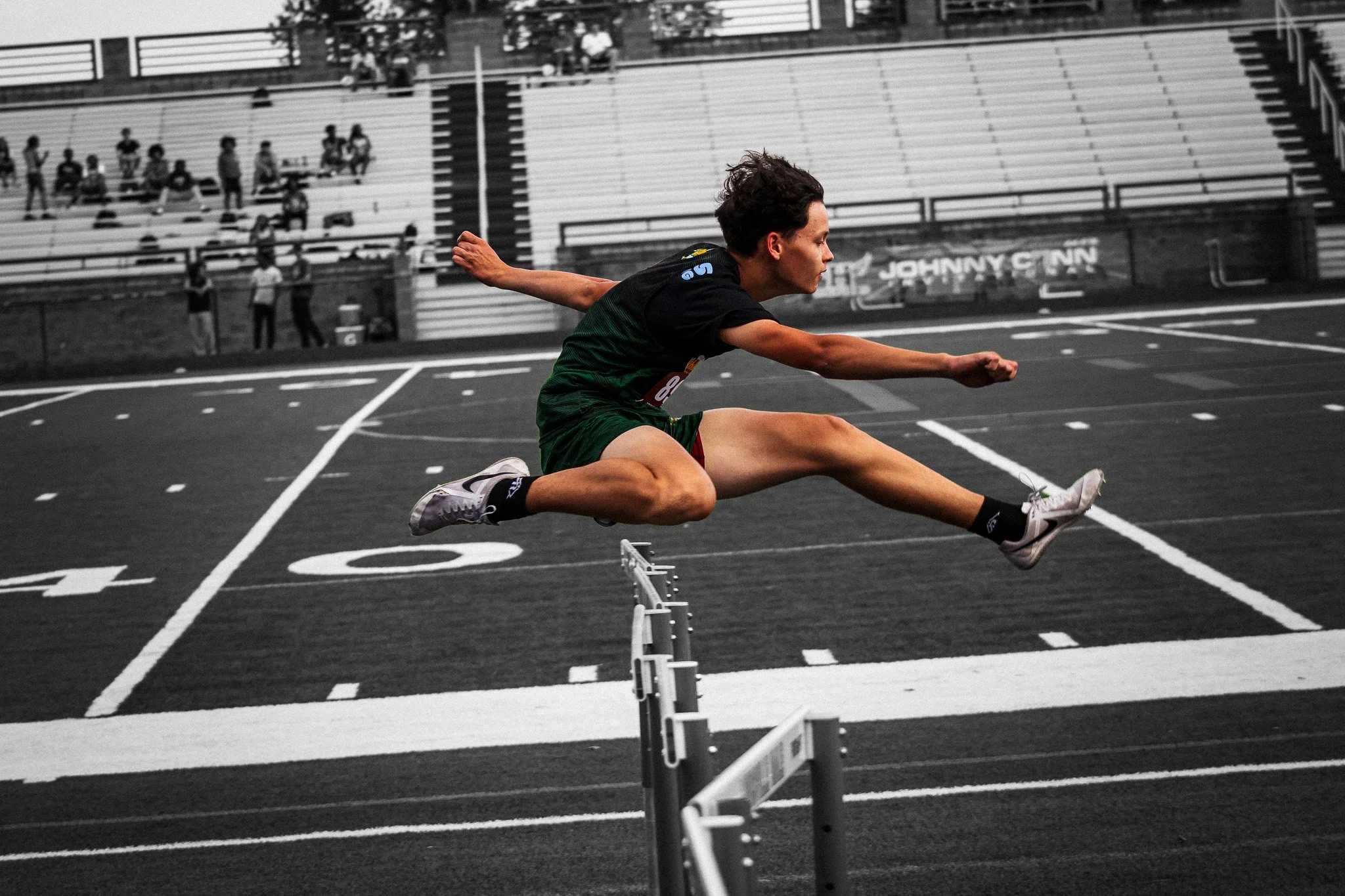 A person with dark hair and light skin is competing in a hurdles race on a track, mid-air jumping over hurdles with arms extended and legs bent. The background shows empty bleachers and a few spectators, with some trees and a sign that reads 'Johnny Cinn'.