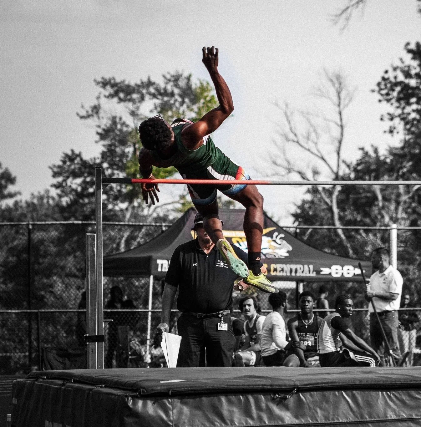 Athlete attempting a high jump over a bar during a track and field competition, with officials and spectators in the background.