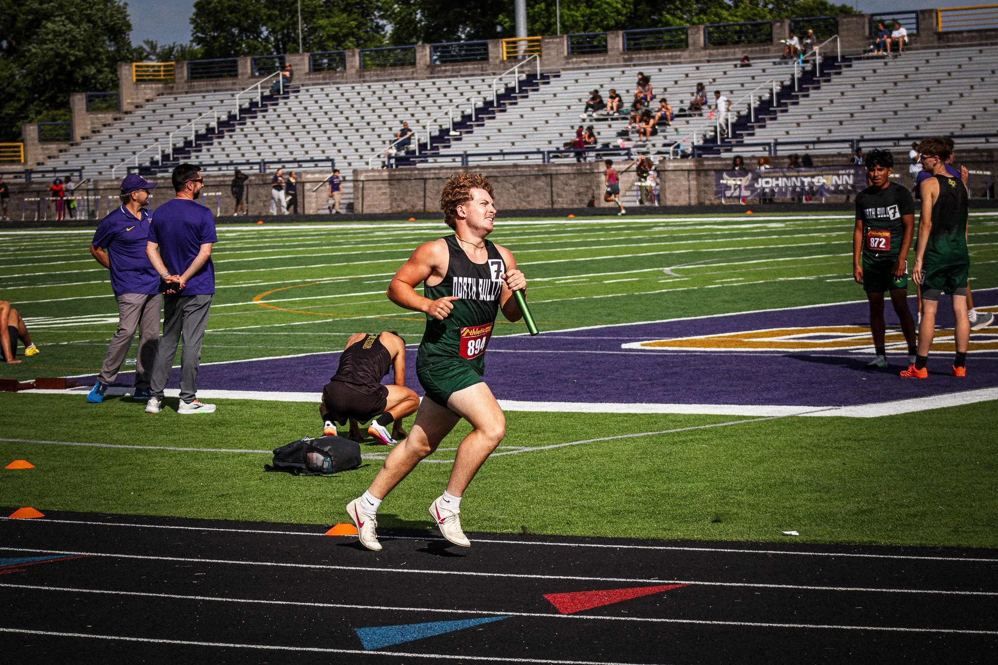 A male athlete in a black and green uniform running on a track with a relay baton, while spectators and other athletes are present in the background at a stadium.