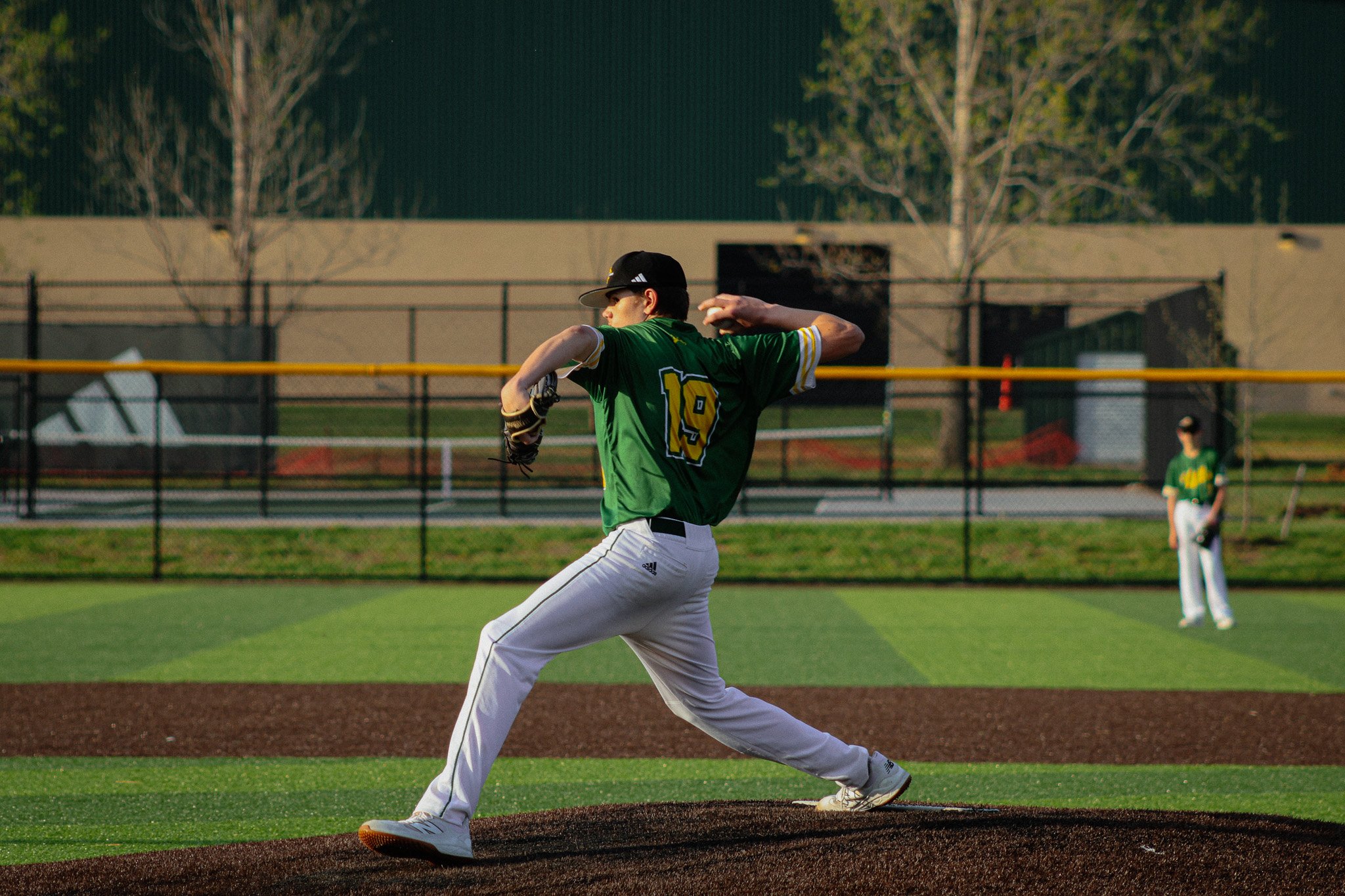 A baseball player in a green jersey and white pants pitching on a field with trees and buildings in the background.