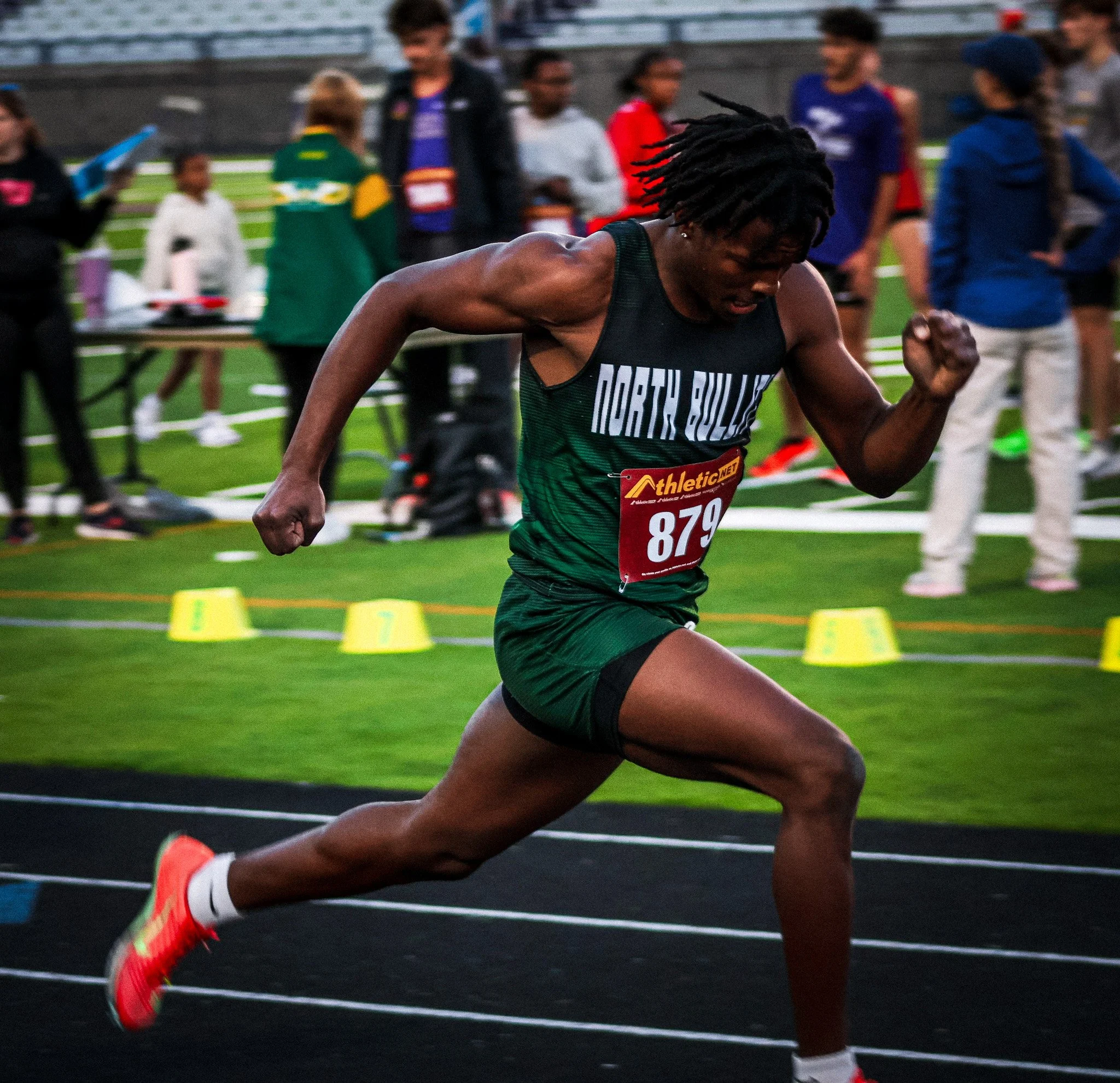 A male athlete with dreadlocks running on a track during a race, with several spectators and participants in the background.