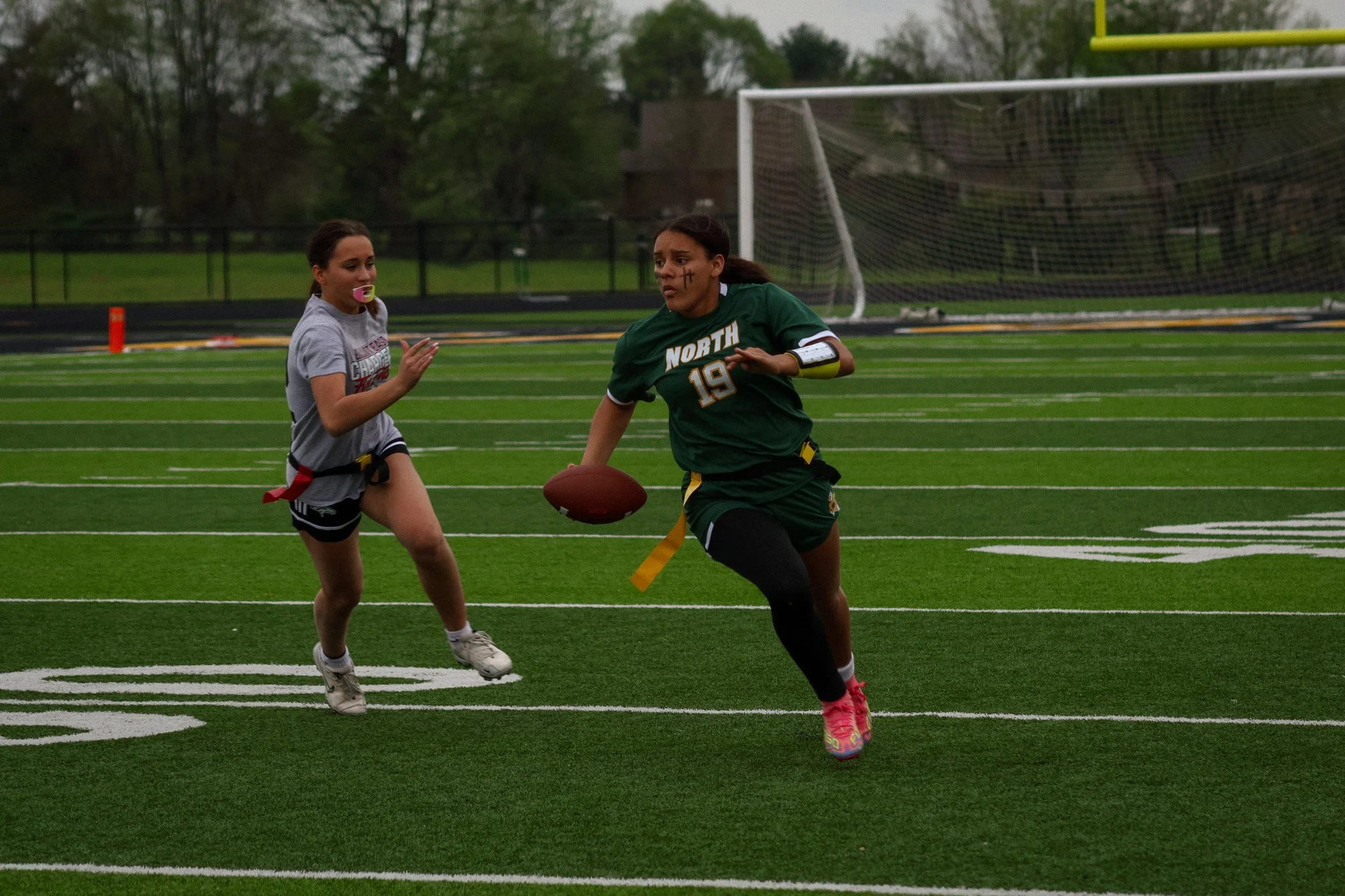 Two women playing football on a field, one in a green uniform with the number 19 and face paint, the other in a gray T-shirt and black shorts, with trees and goalpost in the background.
