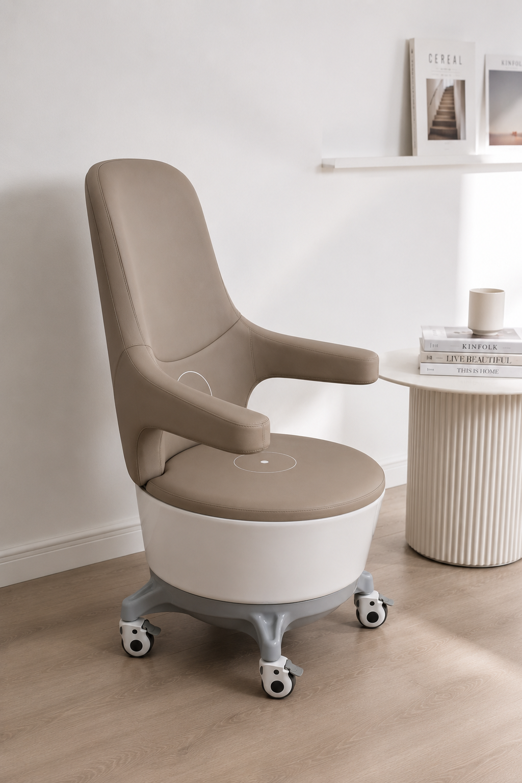 A beige and white office chair with wheels next to a round white side table holding stacked books and a mug, set against a white wall with a shelf of magazines.