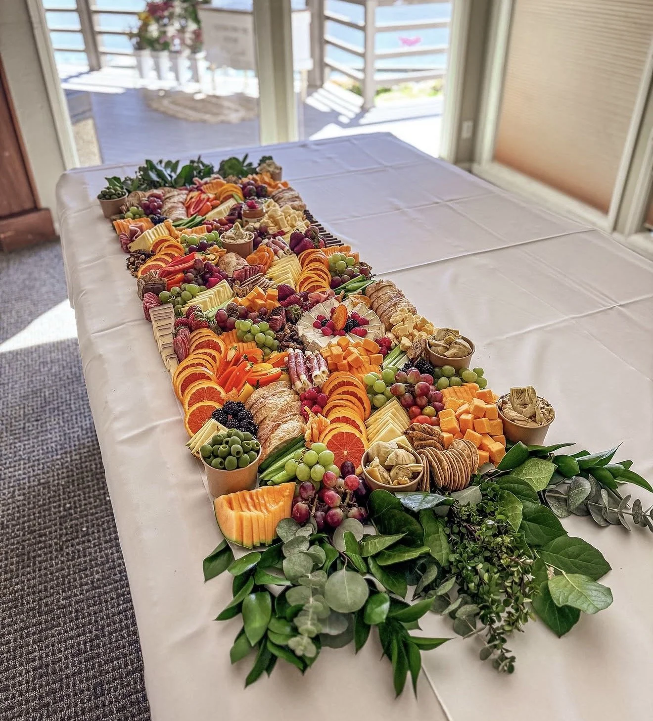 A long platter of assorted fruits, cheeses, crackers, and garnishes on a white tablecloth, with a backdrop of a bright outdoor deck through glass sliding doors.