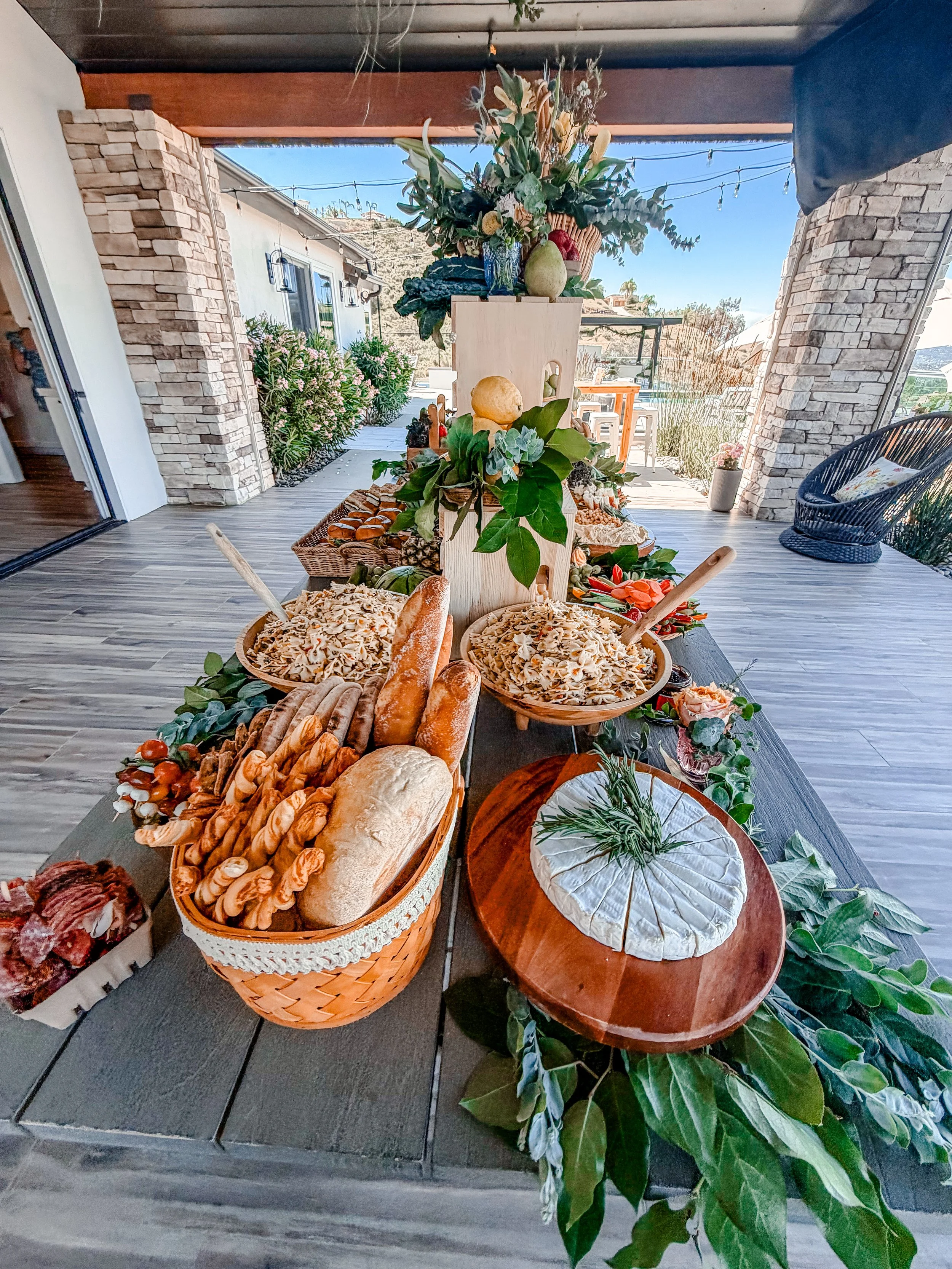 Outdoor table filled with assorted nuts, breads, and snacks, decorated with floral arrangements and greenery, set on a wooden porch with stone walls and string lights in the background.