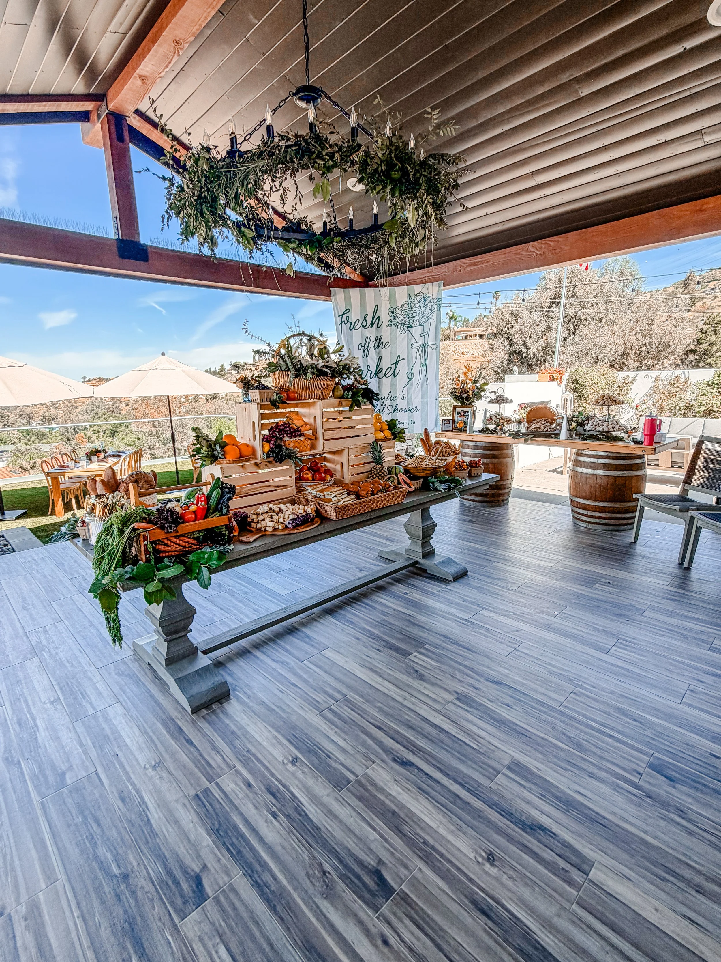 Outdoor market setup with a wooden table displaying assorted fruits and vegetables, decorative flowers, and a banner that reads 'Fresh off the market' with a note about a bridal shower in San Diego, California. There are tables and umbrellas outside, under a covered patio with a chandelier hanging from the ceiling.