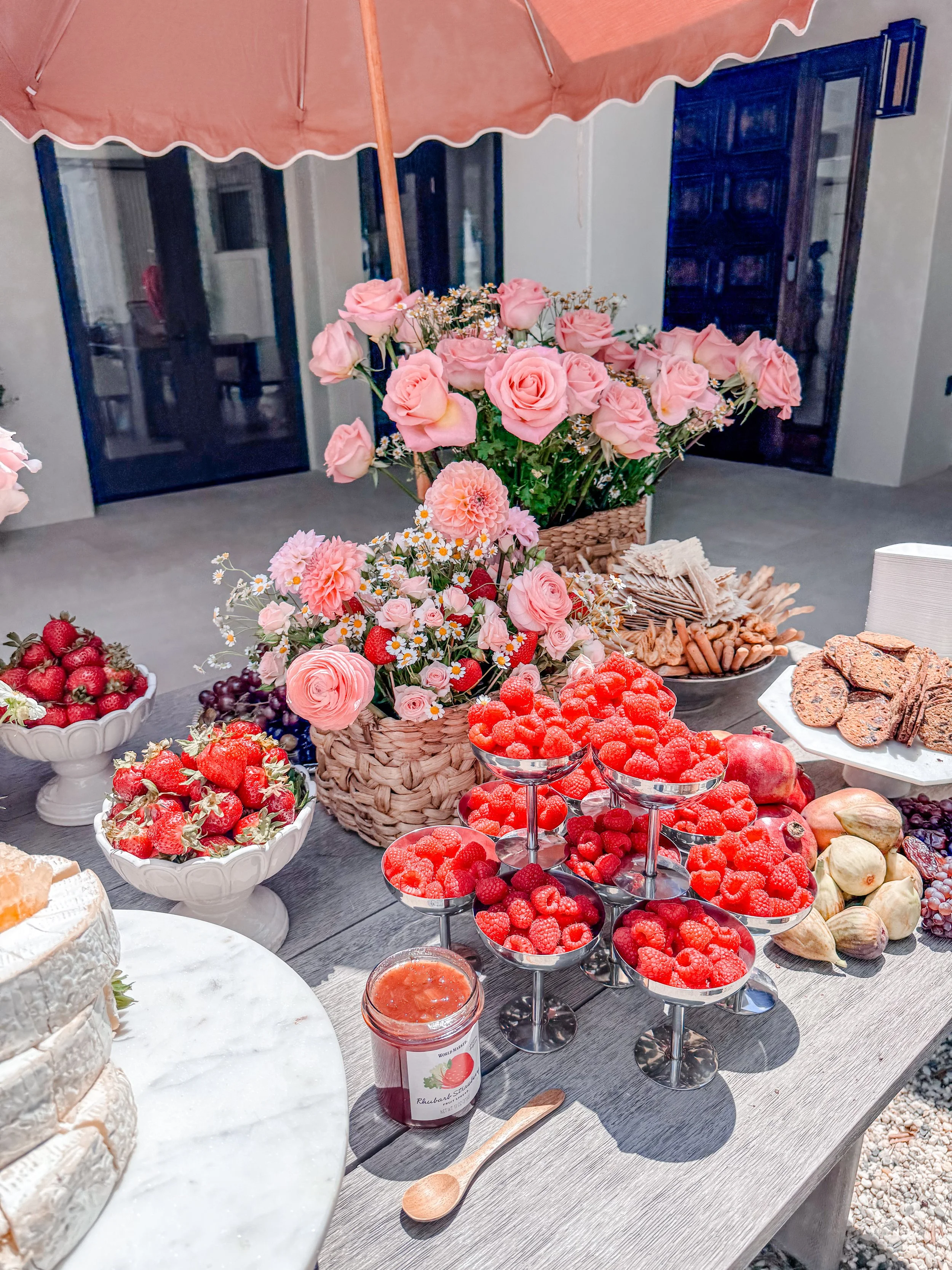 A table decorated with pink roses and daisies in baskets, surrounded by bowls of strawberries, raspberries, and grapes, with cookies and a cake in the background, and an umbrella shading the setup.