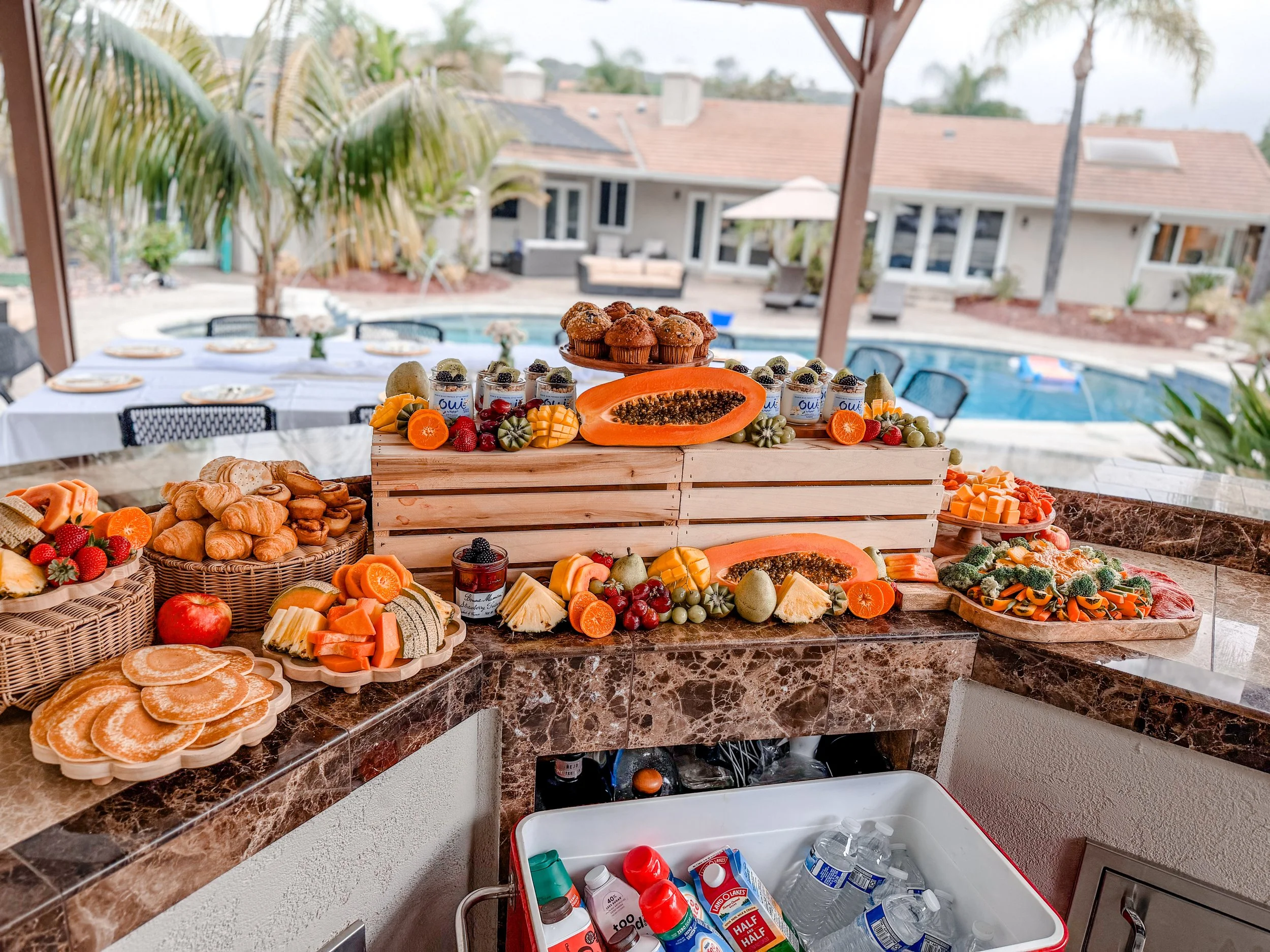 Buffet table with cupcakes, fresh fruit, baked goods, and yogurt with toppings, set in a backyard patio with pool and outdoor furniture in the background.