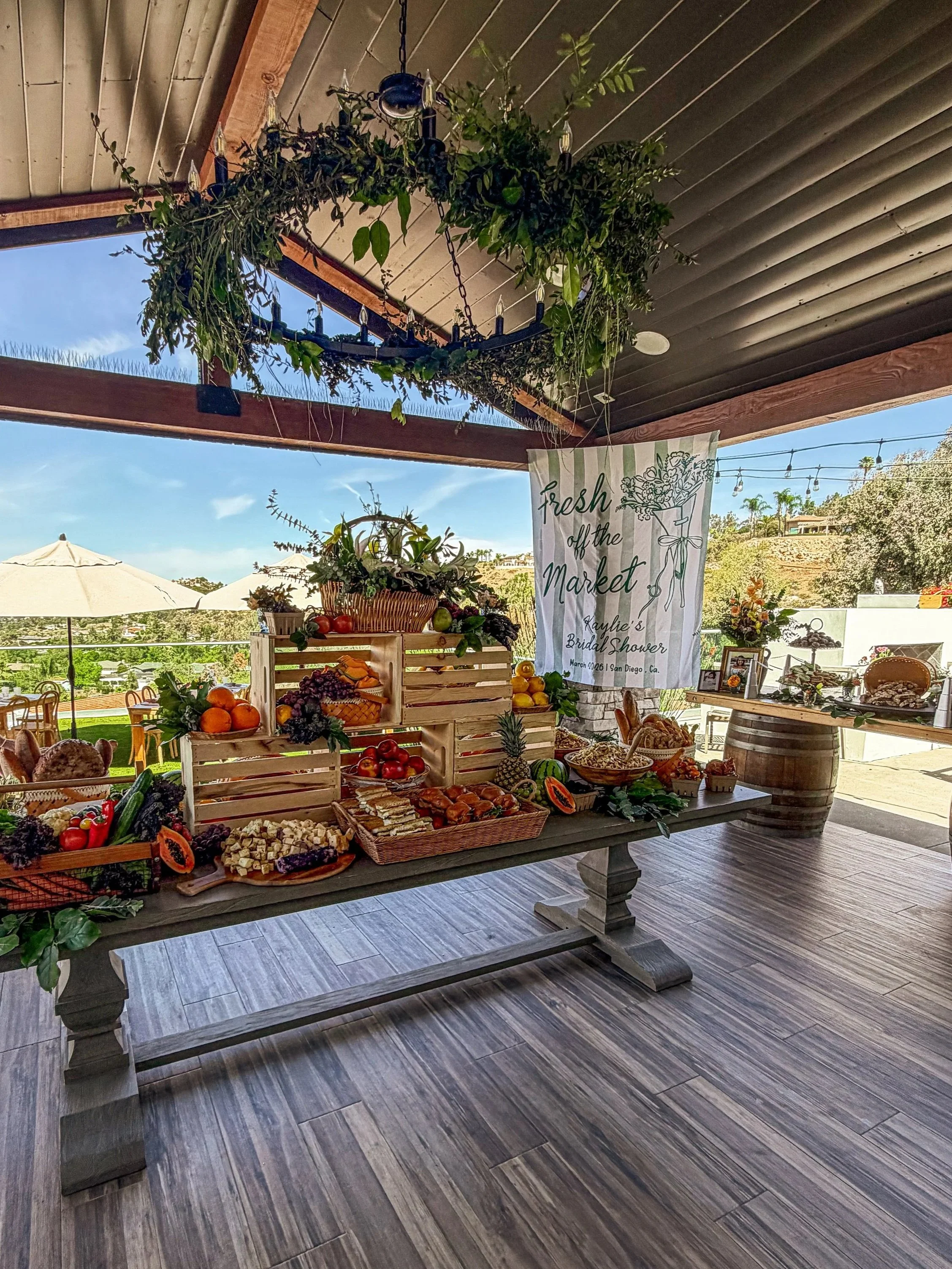 Indoor market display with fruits and vegetables, wooden crates, and a banner that reads 'Fresh off the Market Kaylie's Bridal Shower March 2021 San Diego, Ca.' A chandelier made of greenery hangs from the ceiling. Outdoor scenery with umbrellas and trees is visible through open windows.