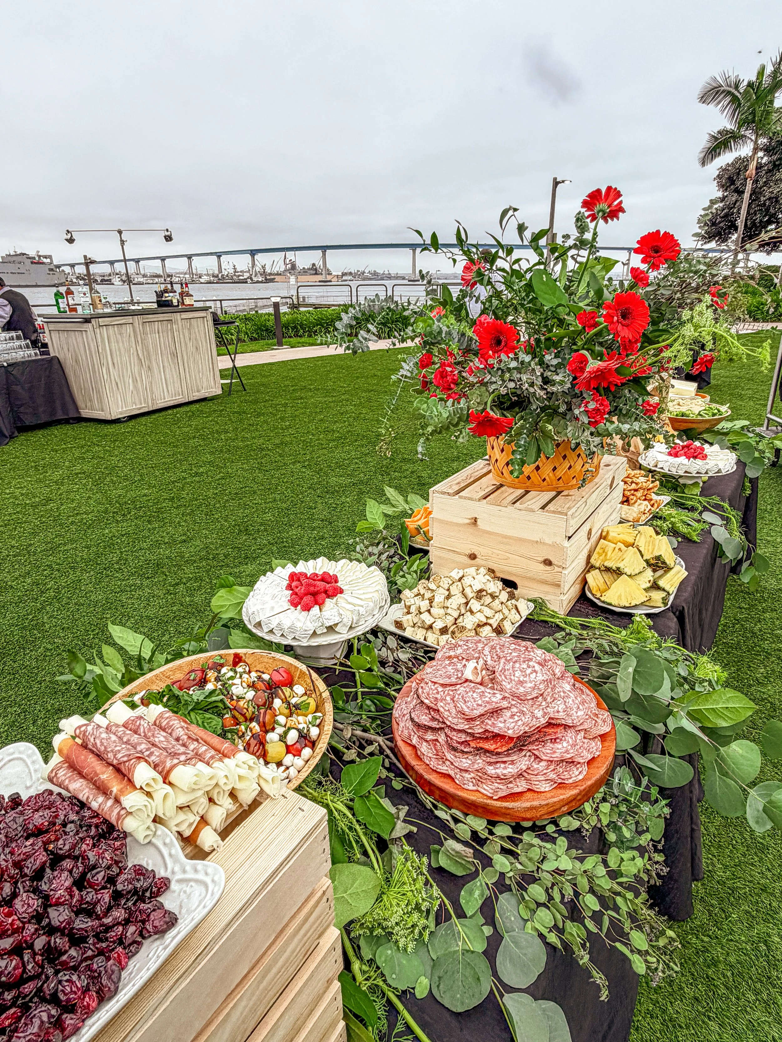A charcuterie buffet table with sliced meats, cheese, and berries, decorated with a large bouquet of red flowers, set outdoors near water and a bridge.