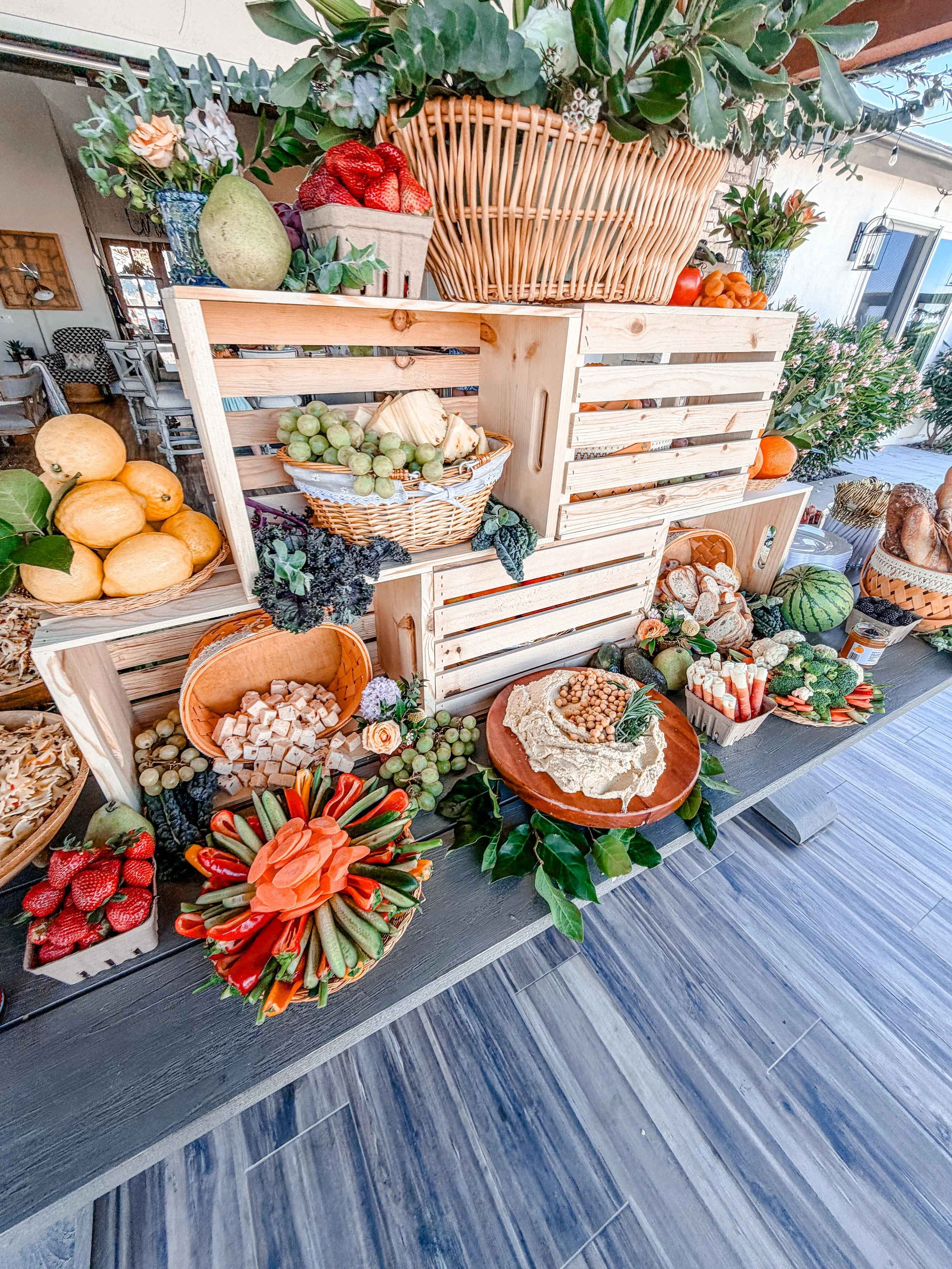 Display of fresh fruits, vegetables, and flowers arranged in wooden crates and bowls, including strawberries, grapes, watermelon, pumpkins, and leafy greens, on a wooden table with a floral backdrop.