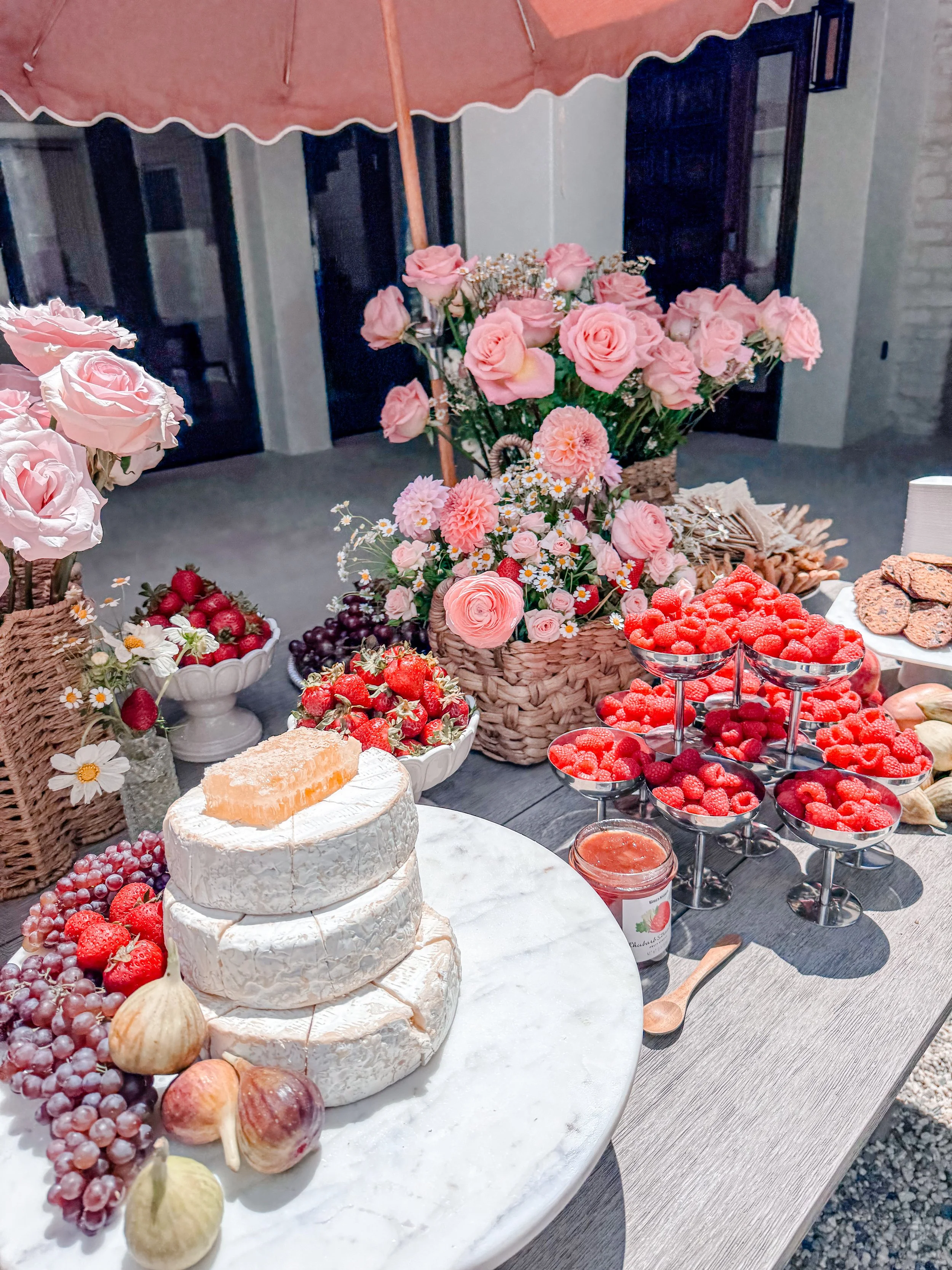 A dessert table with a tiered cake, fresh strawberries and grapes, and pink roses in baskets, set on a white marble surface with jars of jam and a spread of crackers, under a pink patio umbrella.