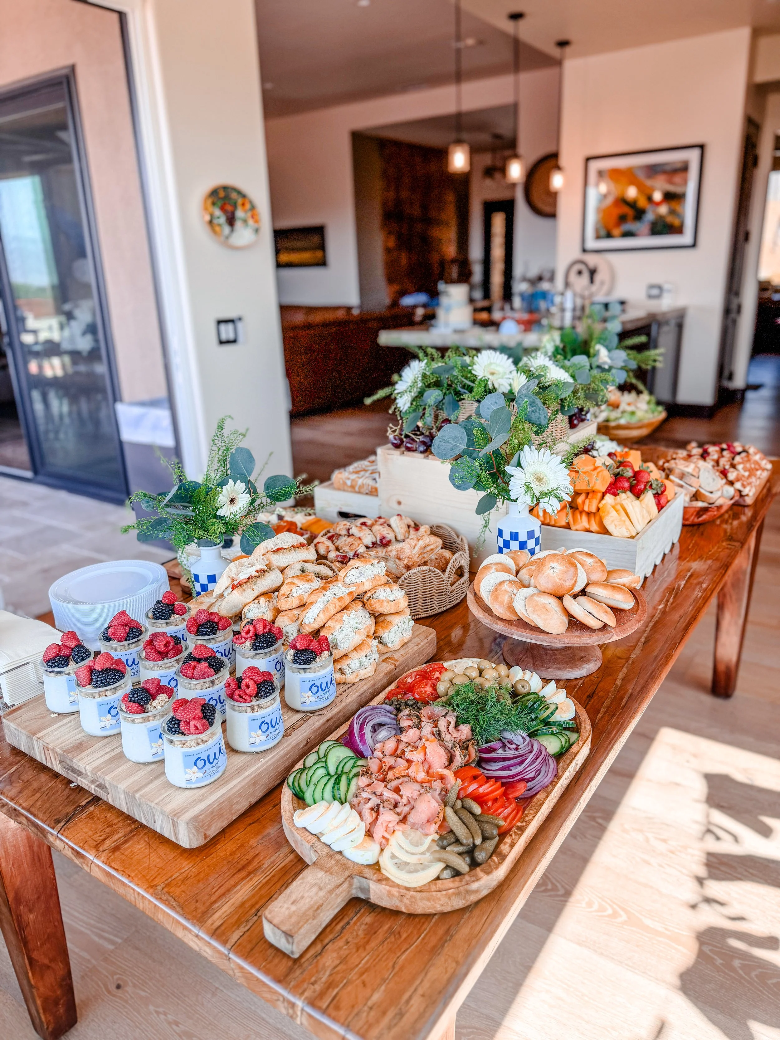 A buffet table with various appetizers, cheeses, crackers, fruits, and vegetable platters, decorated with floral arrangements, in a cozy indoor setting.