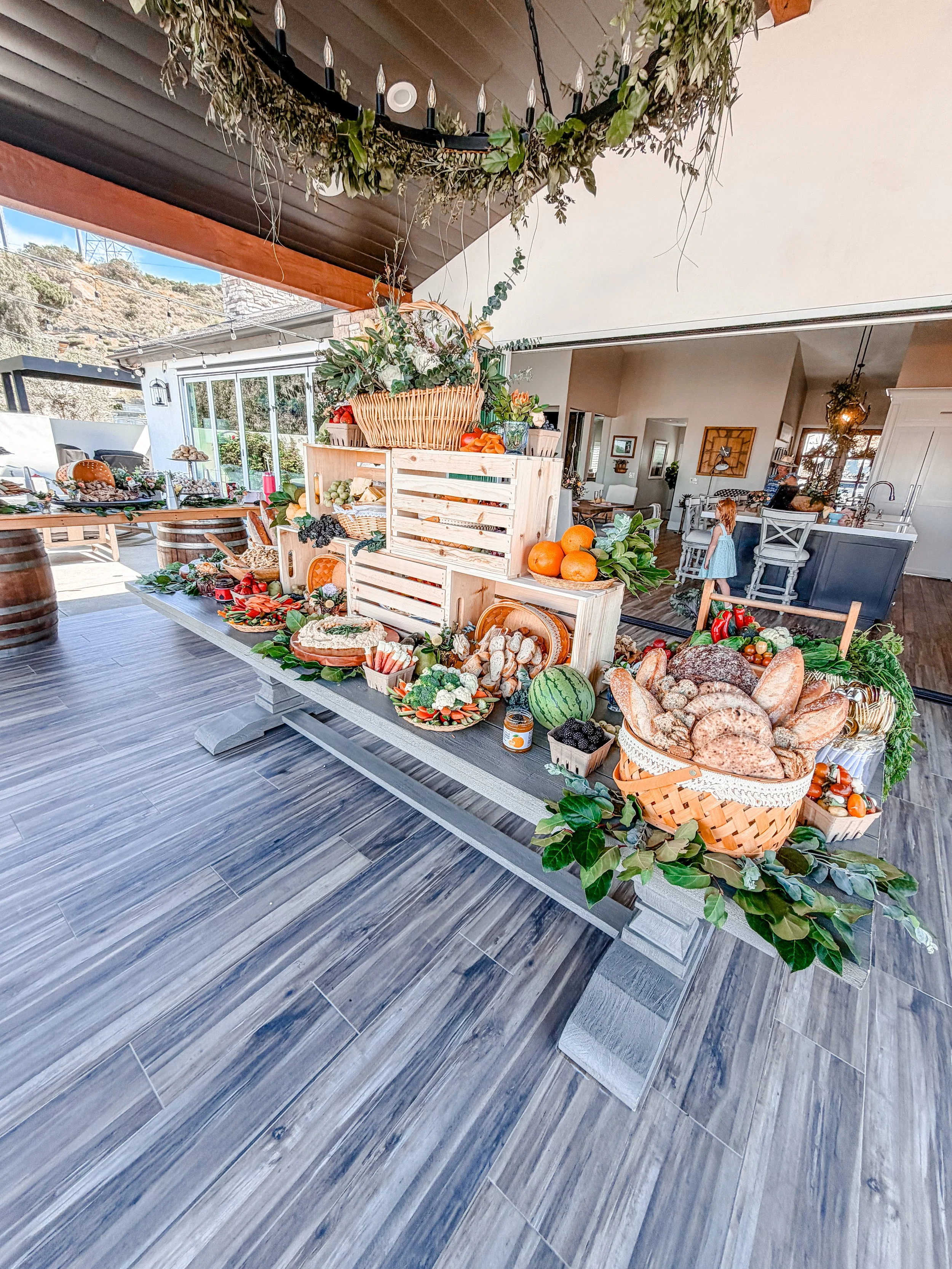 Indoor market display with fresh fruits, vegetables, bread, and decorative greenery, set on a wooden table with a rustic background and tiled flooring.