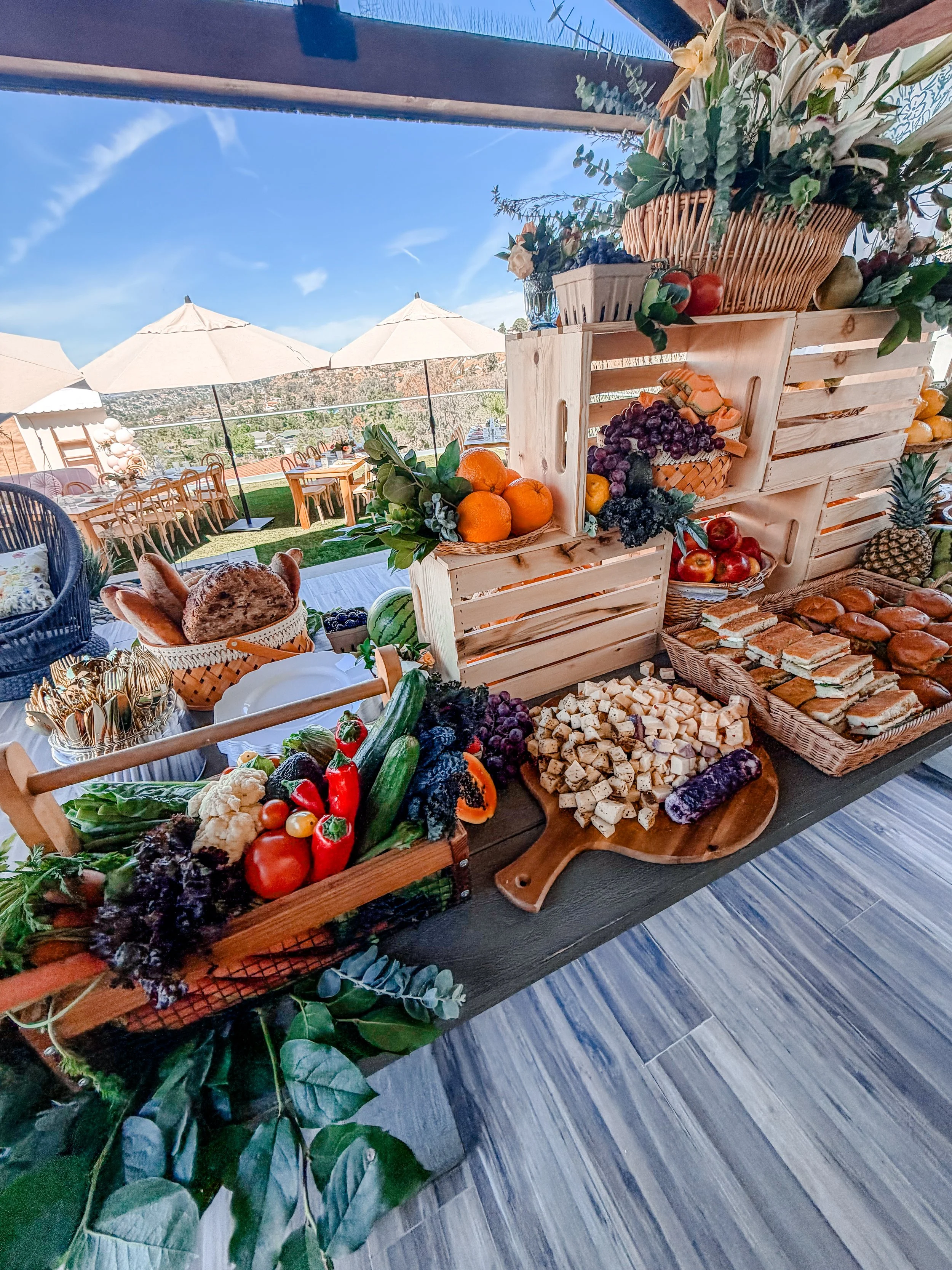 A table filled with various fruits, vegetables, and bread, with a backdrop of outdoor seating and umbrellas on a sunny day.