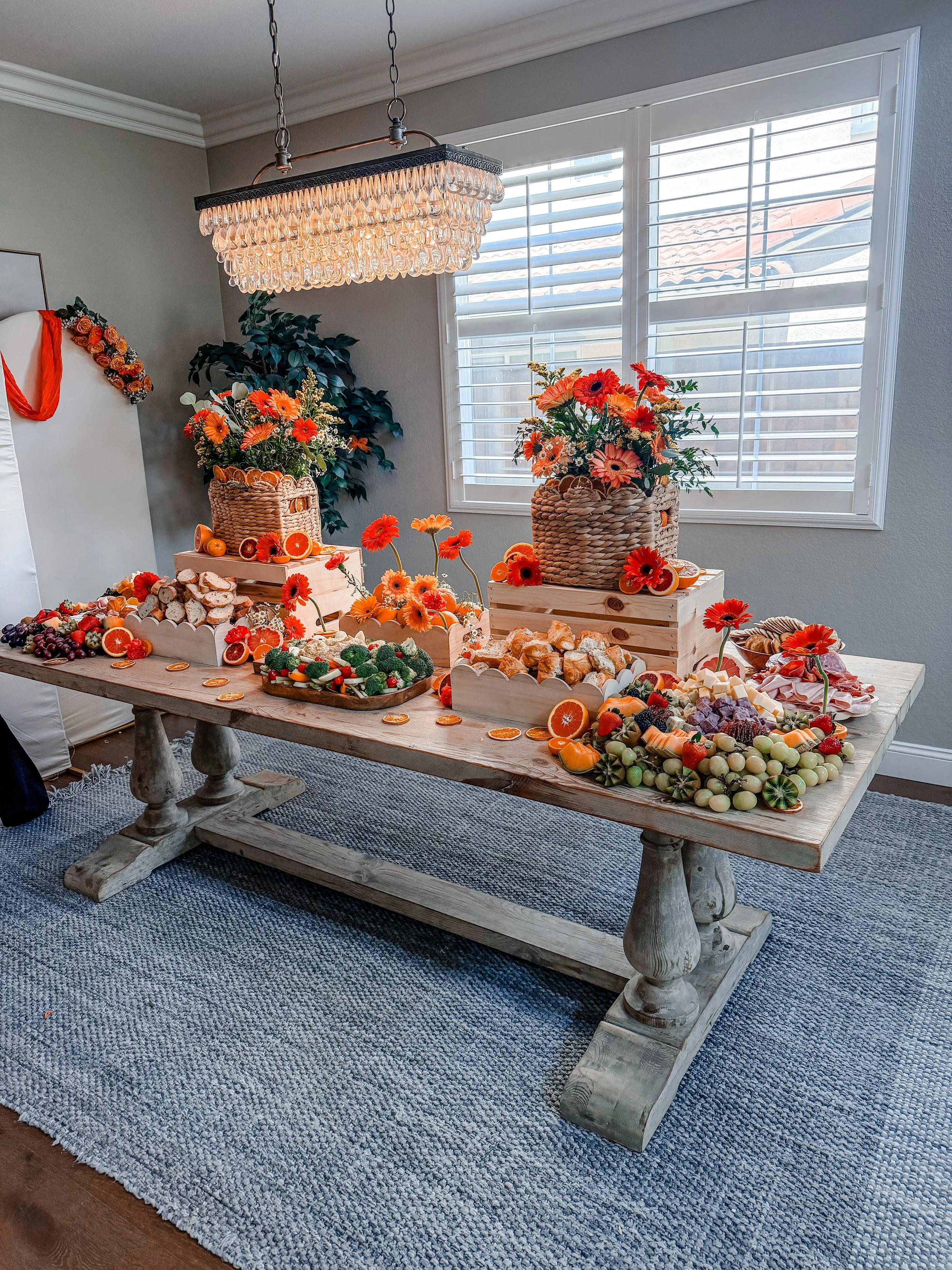 A large dining table with a rustic wooden surface and sturdy legs, decorated with an abundance of bright orange and red flowers, citrus fruits, and assorted snacks. There are two large woven baskets filled with flowers, as well as wooden crates and trays holding various colorful fruits, vegetables, cheeses, and pastries. The room has a window with plantation shutters and a decorative chandelier hanging above the table, with a blue and gray area rug beneath.