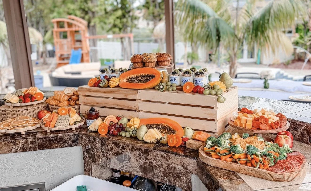 A buffet table filled with assorted fruits, vegetables, and baked goods, including papayas, strawberries, grapes, muffins, and cheese cubes, set outdoors with trees and a backyard playset in the background.