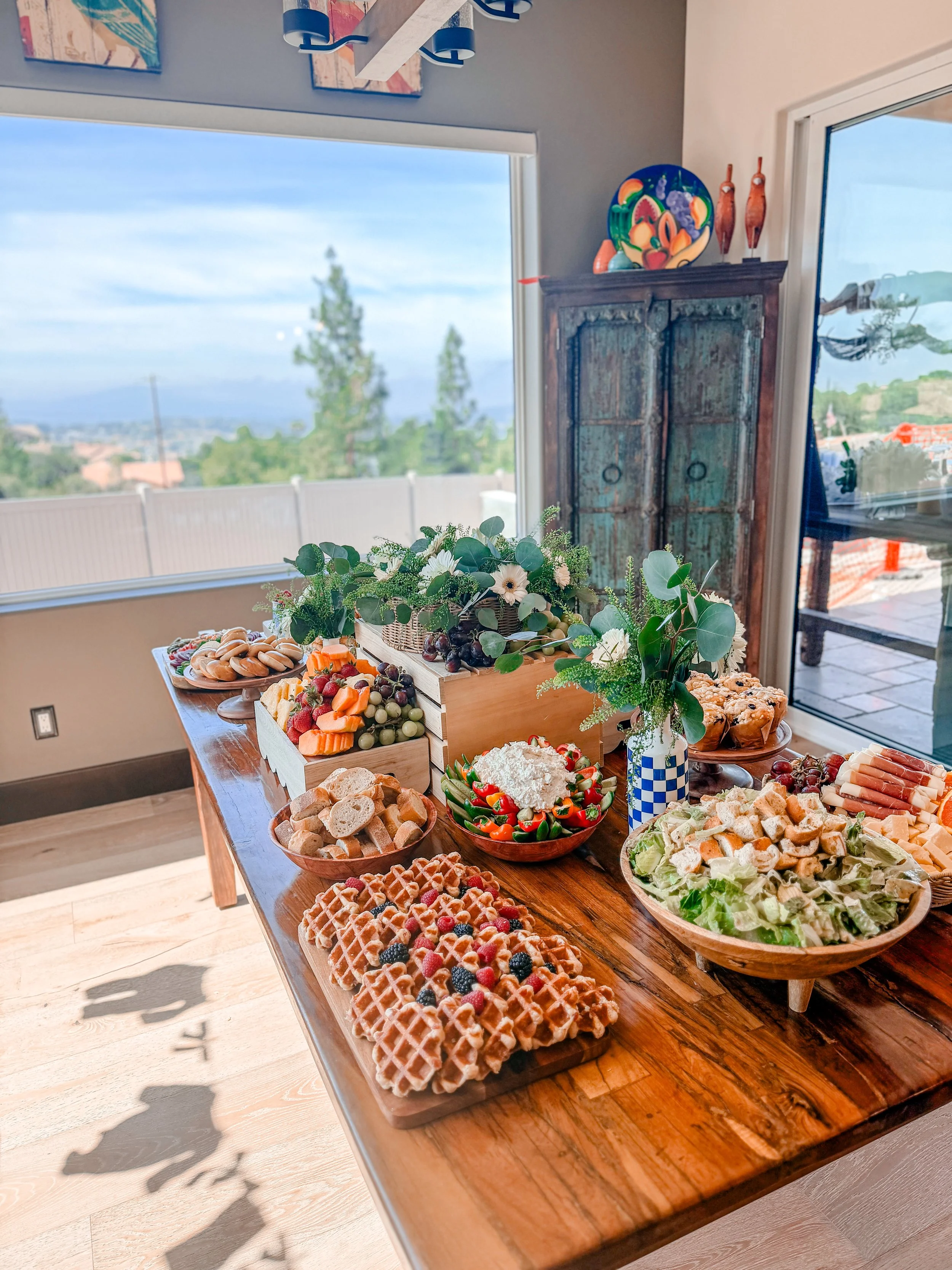 A table with assorted breakfast foods including waffles, fruit, bread, and salads, set in a room with large windows showing a sunny outdoor scene.