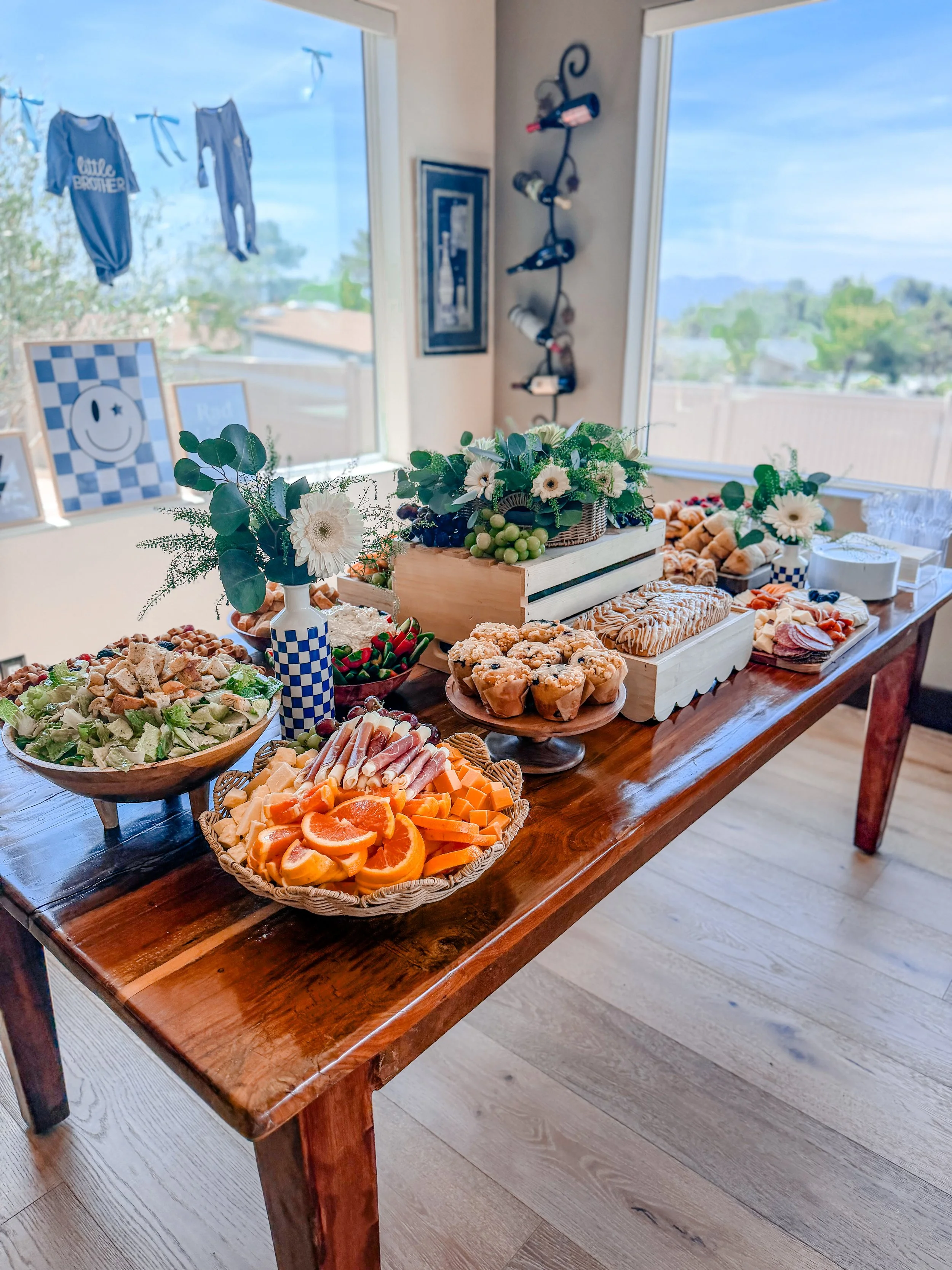 A wooden table set with various appetizers, including sliced oranges, cheese, sliced meats, bread, grapes, and a salad, decorated with flowers and greenery in vases.