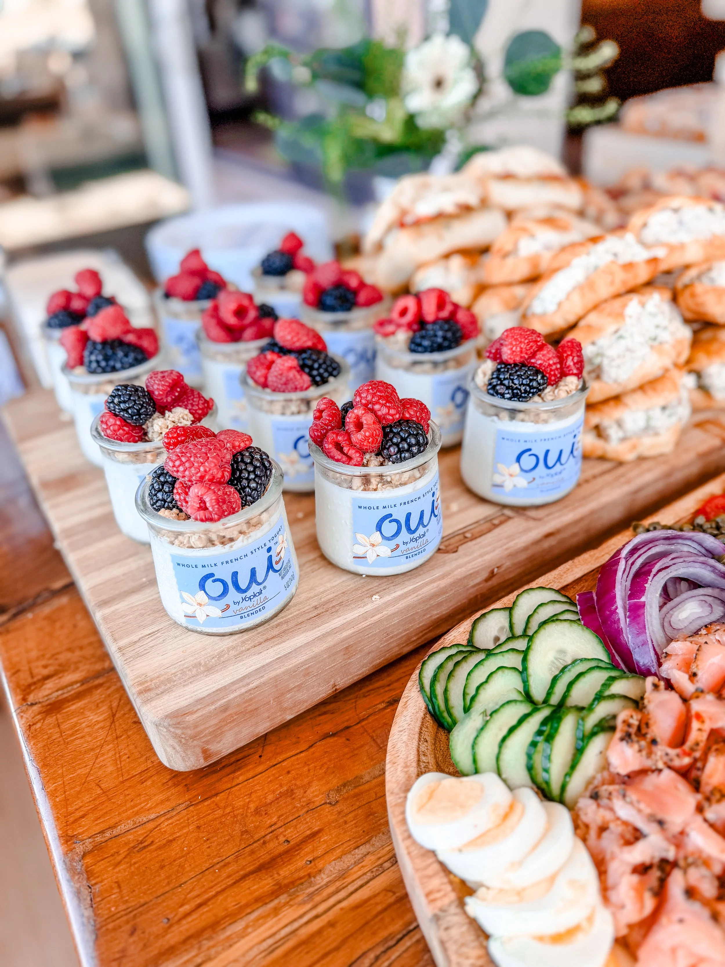 Small glass jars of vanilla yogurt topped with mixed berries, arranged on a wooden board at a buffet. To the right, sliced cucumbers, red onions, and a plate of smoked salmon with sliced eggs are also visible.