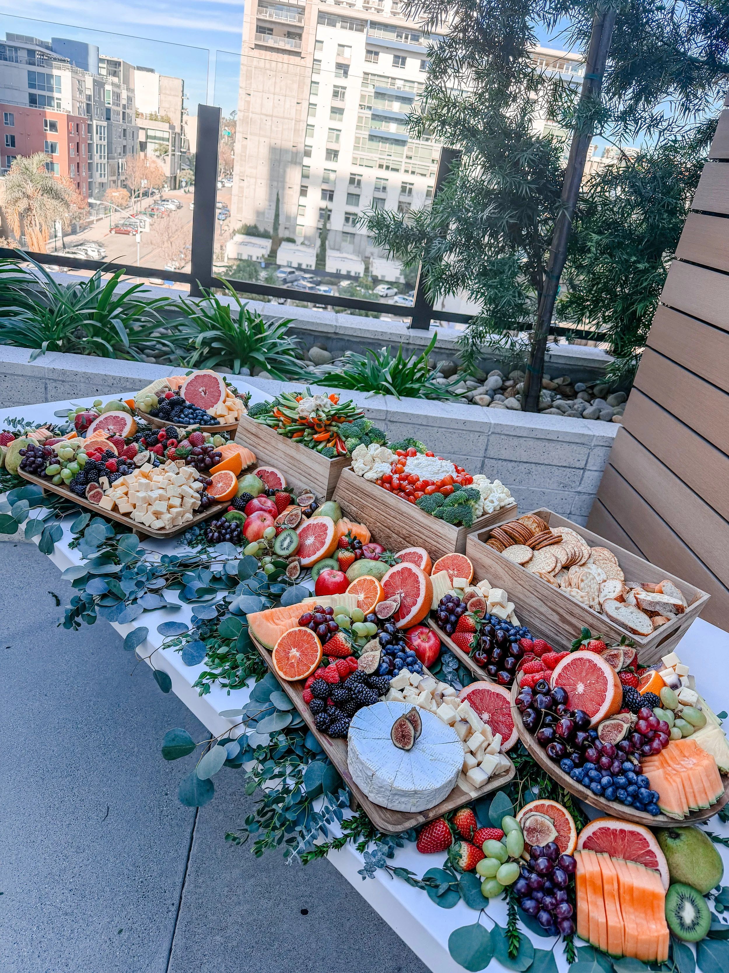 An outdoor table with an assortment of fresh fruits and cheeses, set against a cityscape background with buildings and greenery.