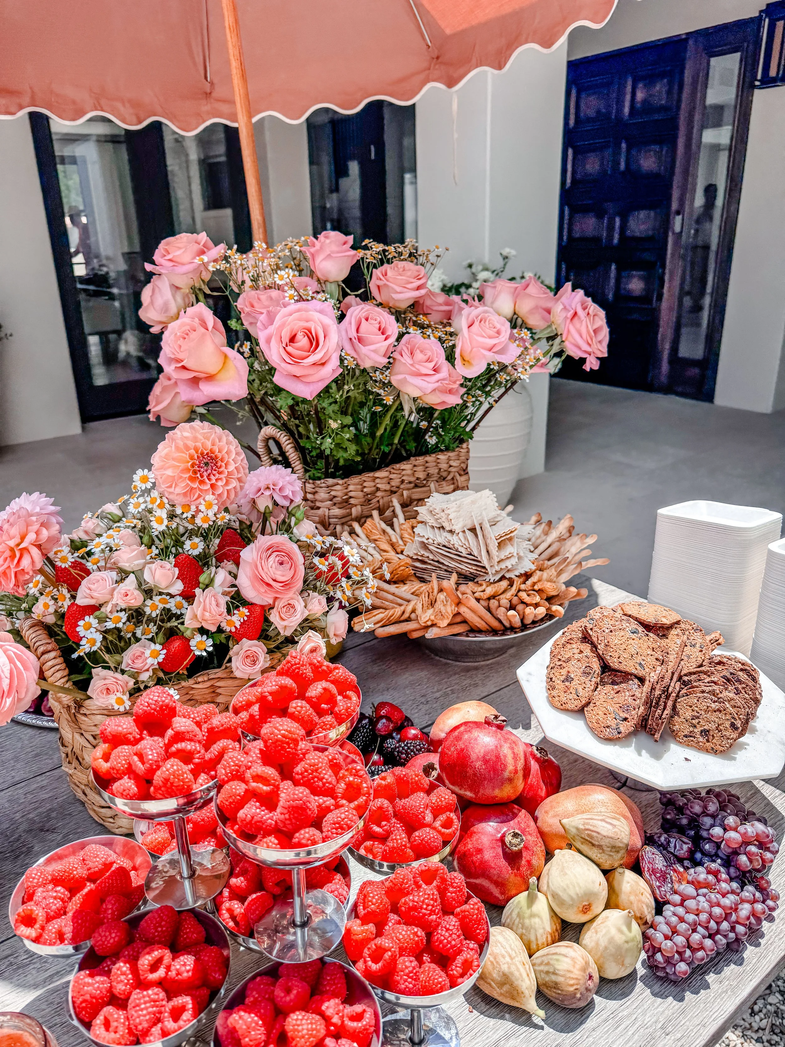 A table of fresh strawberries, raspberries, blackberries, and grapes with potted pink roses and other flowers, arranged for a celebration or gathering.