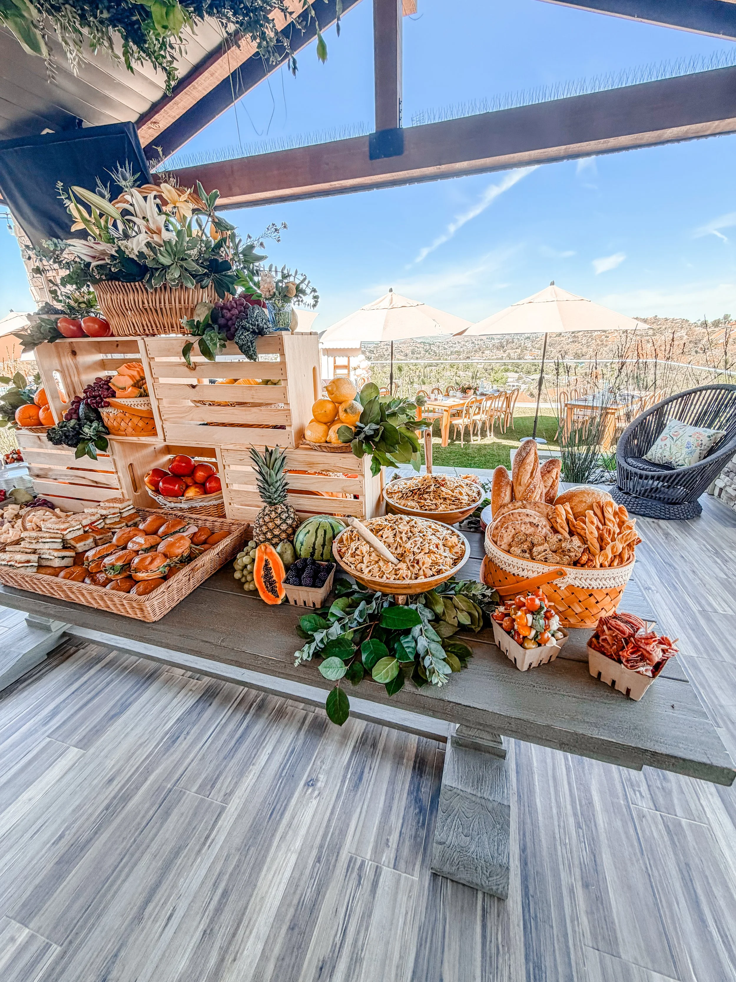 Outdoor buffet table with various fruits, breads, and snacks under a covered patio, with tables, chairs, and umbrellas overlooking a scenic landscape in the background.